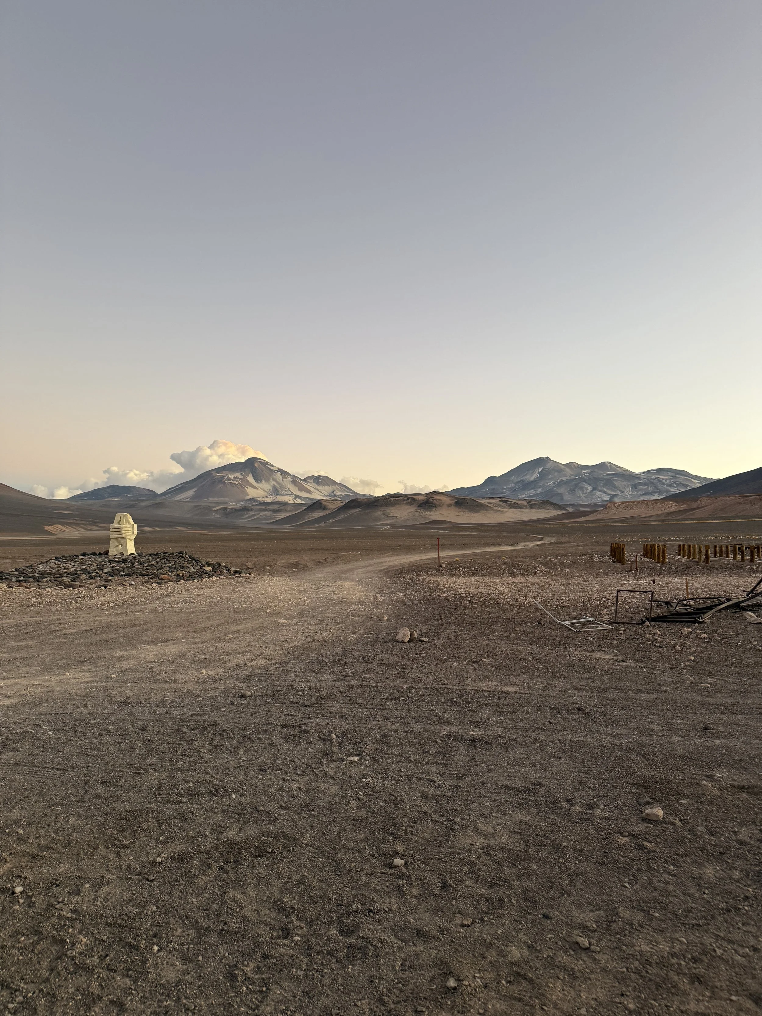 A wide, barren desert landscape with a dirt road, distant mountains, and volcanic peaks in the background, under a clear sky.
