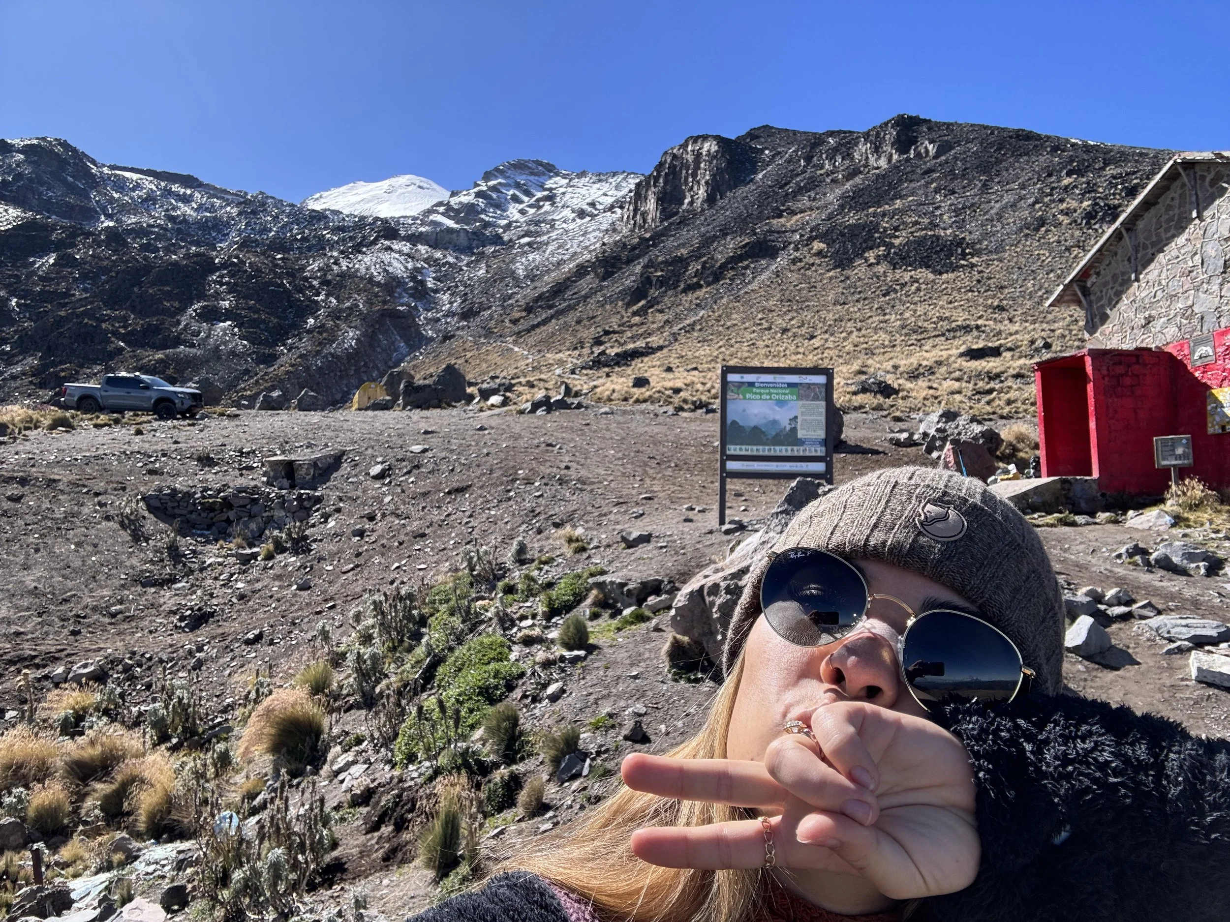 A woman taking a selfie outdoors in a mountainous area with snow-capped peaks and clear blue sky, wearing sunglasses, a gray beanie, and a black jacket.