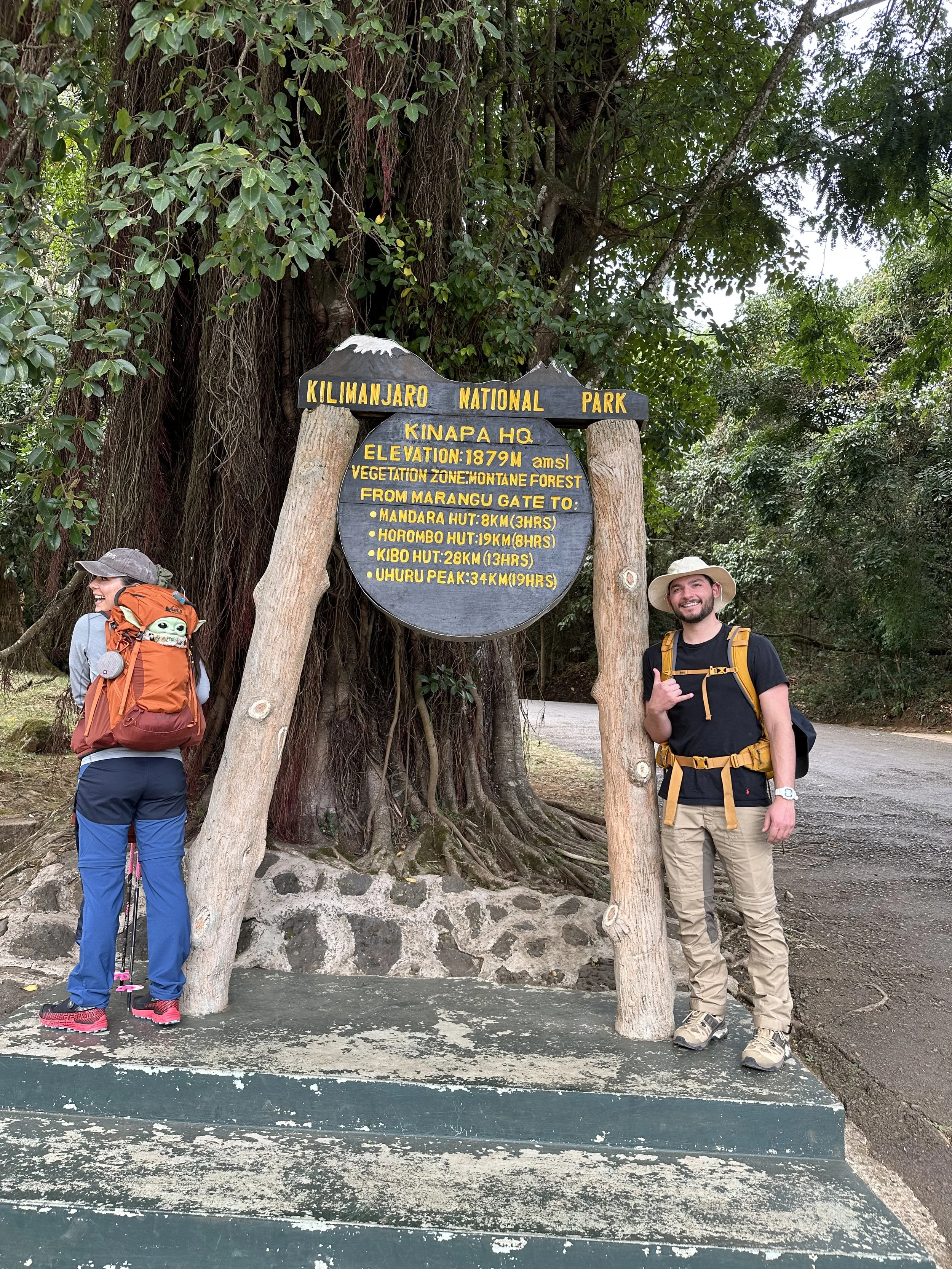Two hikers with backpacks standing next to a wooden sign at Kilimanjaro National Park. One person is facing away from the camera, wearing a gray hat, orange backpack, blue pants, and pink shoes. The other person is facing the camera, wearing a wide-b