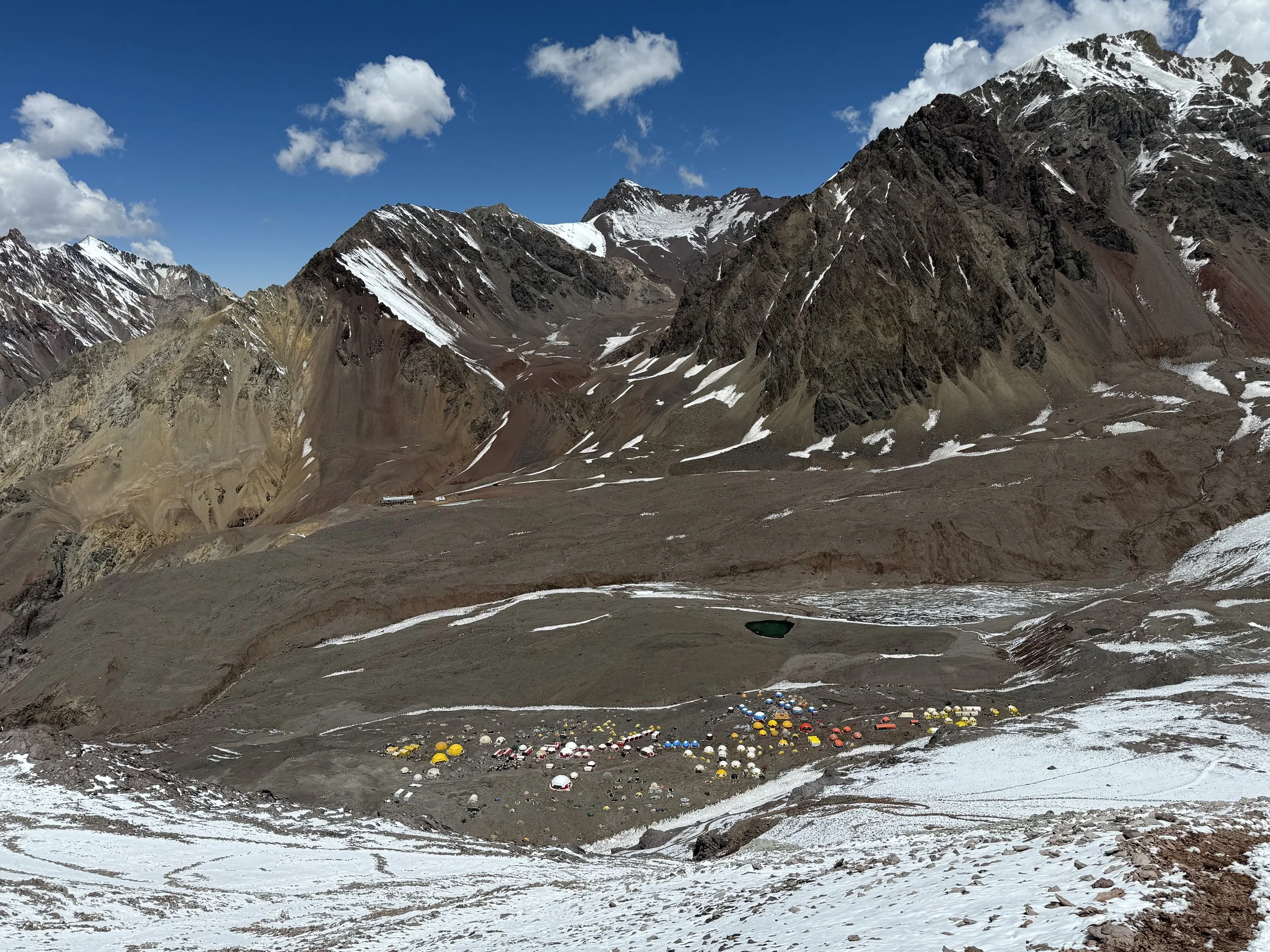 A high-altitude mountain landscape with rugged peaks, some snow-capped, and a valley with a camping site featuring numerous colorful tents and structures.
