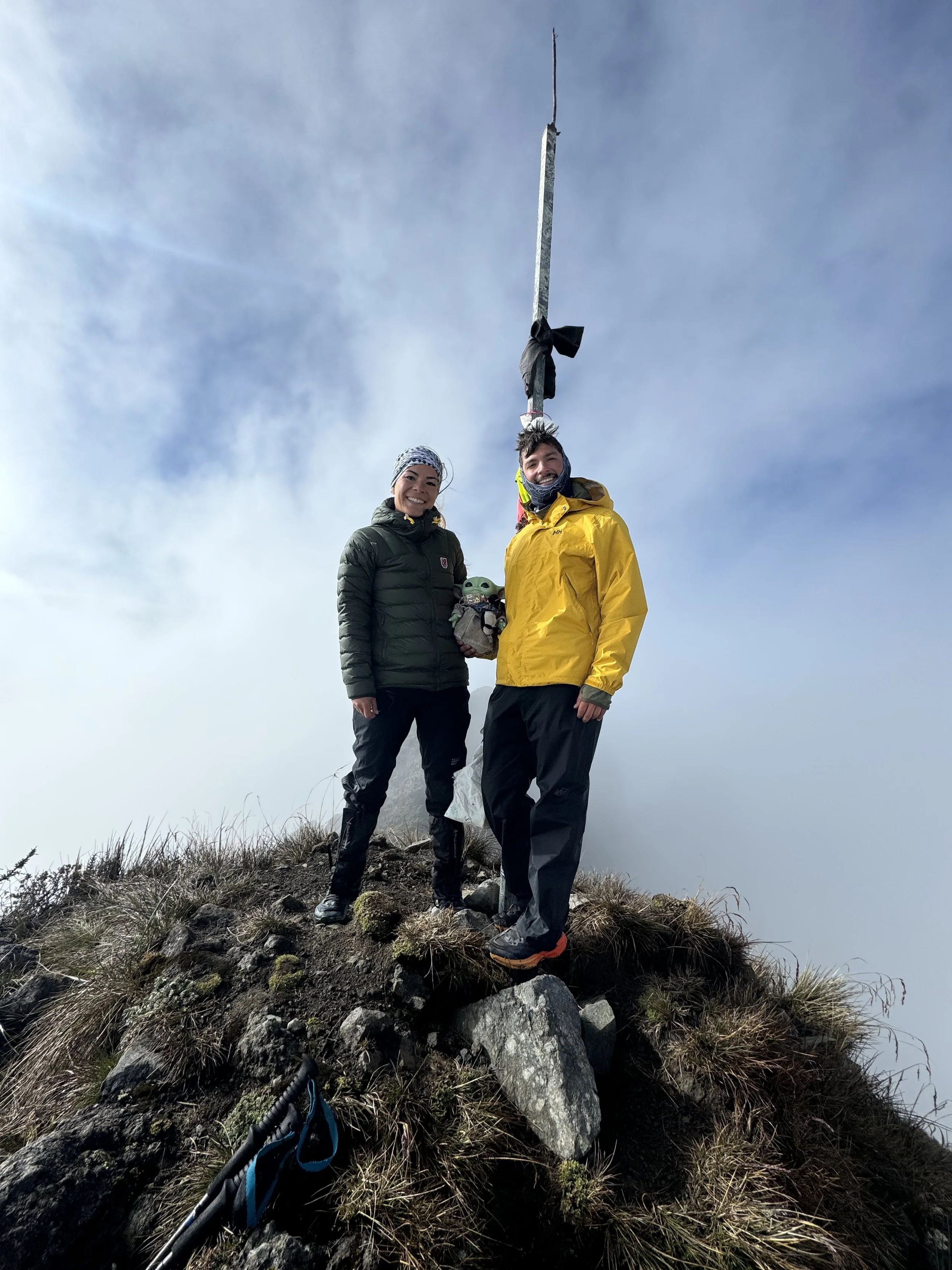 Two hikers stand on top of a mountain peak next to a tall metal pole with a black cloth tied around it. They are dressed in outdoor gear, with one in a black jacket and the other in a yellow jacket. The background shows a cloudy sky.