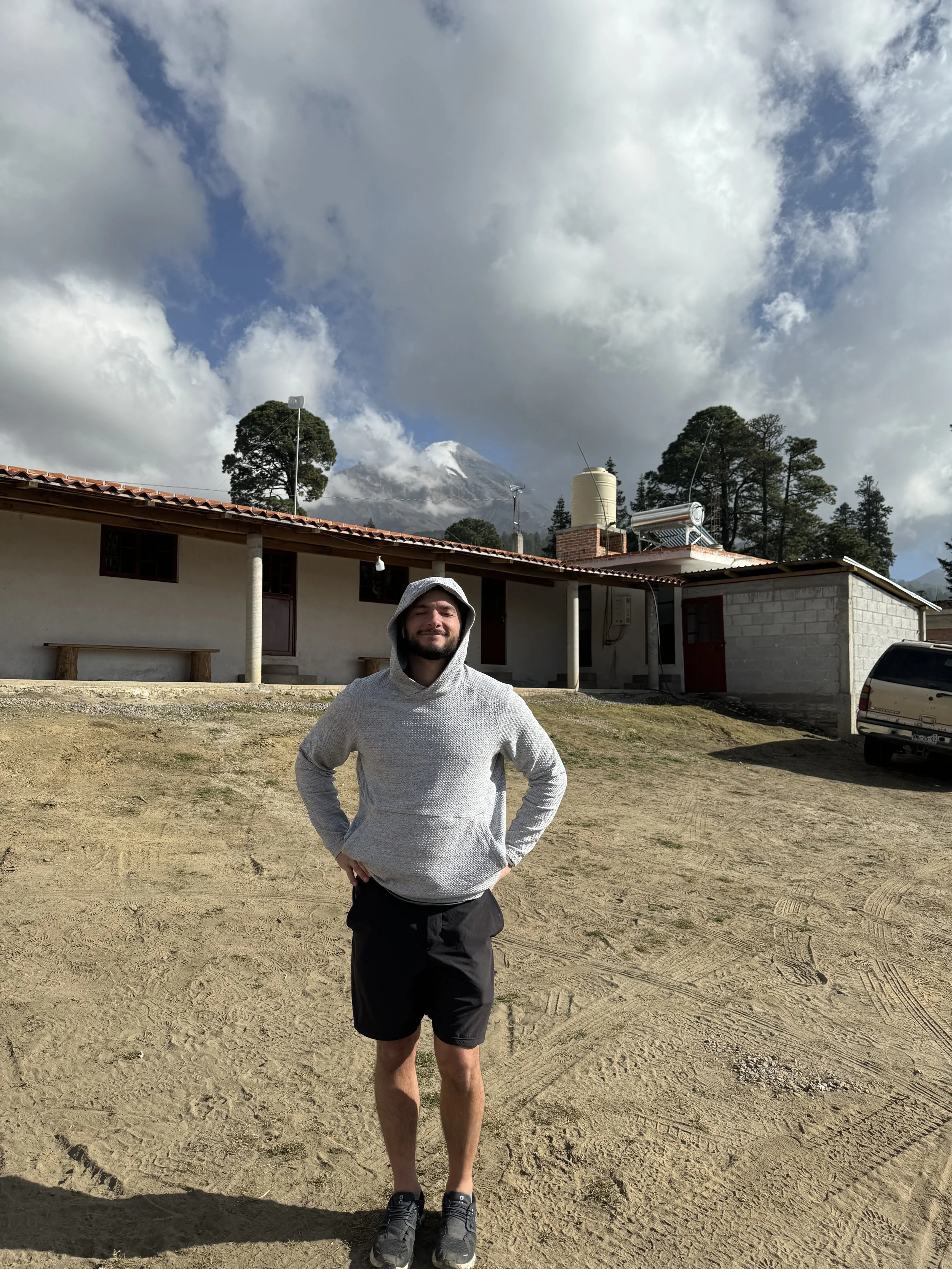 A man stands outdoors wearing a gray hoodie and black shorts, with a building and trees behind him, and snow-capped mountains and a partly cloudy sky in the background.