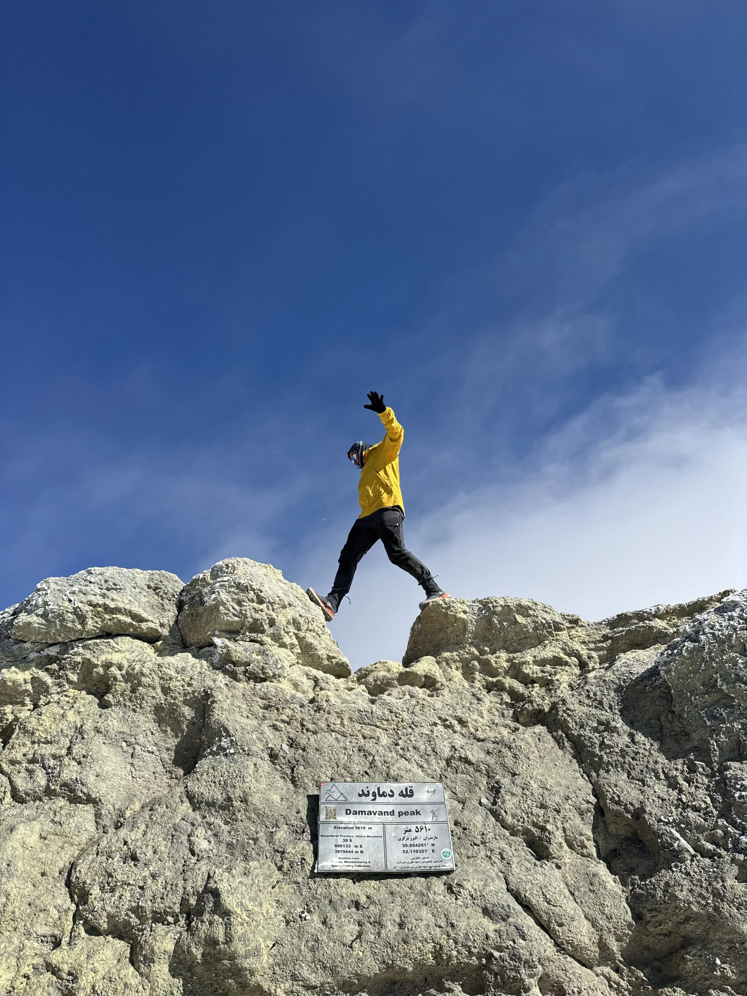 Person in a yellow jacket and black pants walking on rocks at Damavand Peak, high altitude mountain, under a clear blue sky, waving.