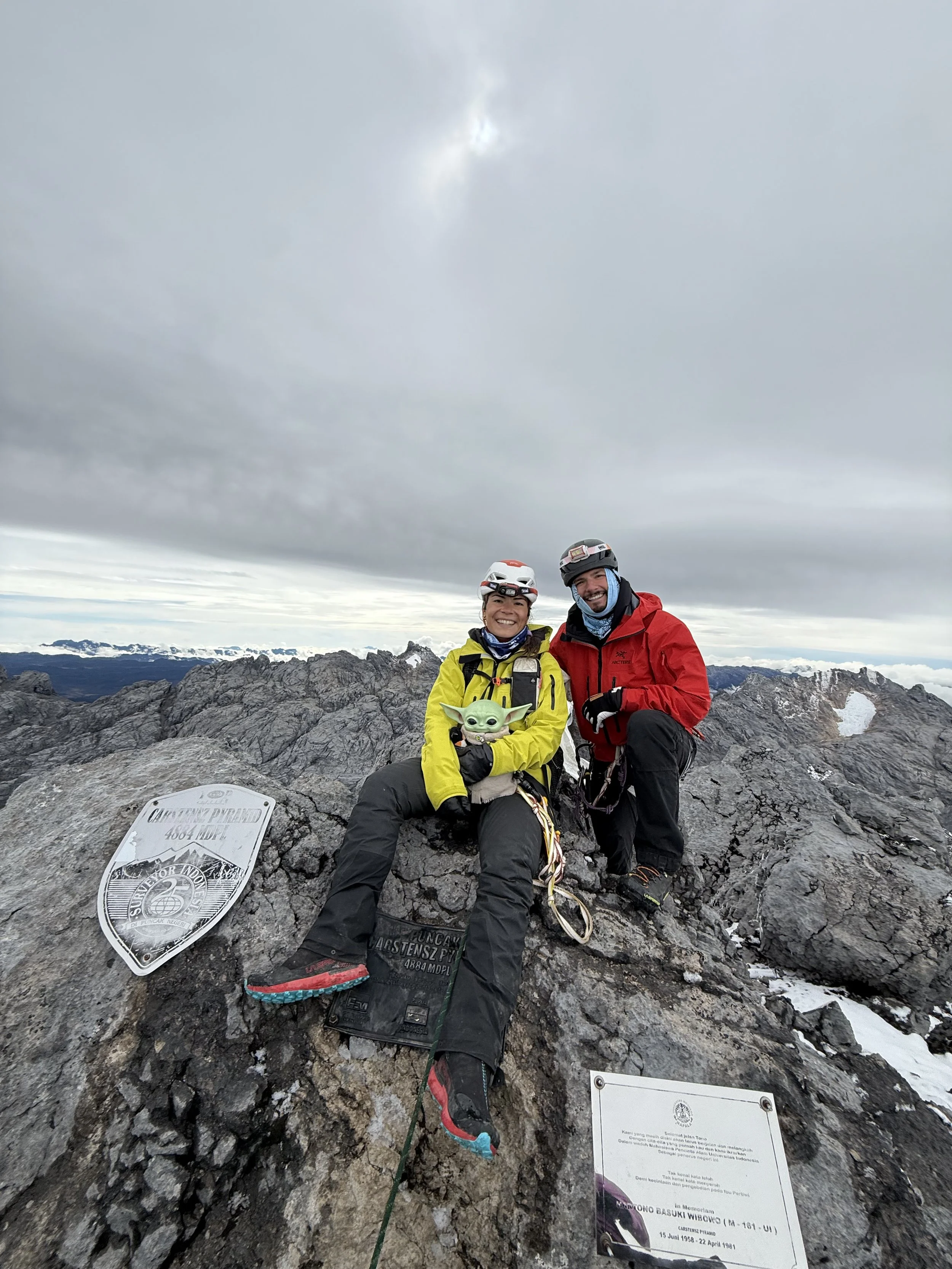 Two climbers at the summit of a mountain, smiling and holding a small green Baby Yoda toy, with mountain peaks in the background and a cloudy sky overhead.
