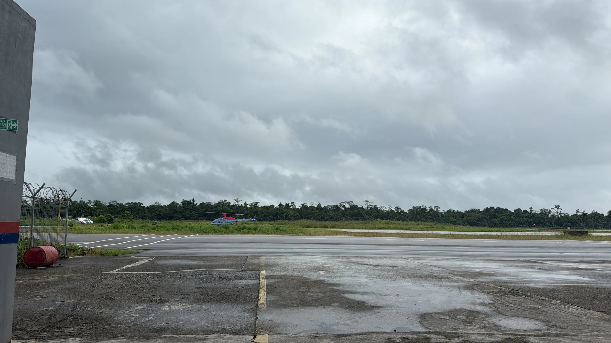 Airport tarmac with puddles, a fence, and two helicopters parked on the grass, under a cloudy sky.