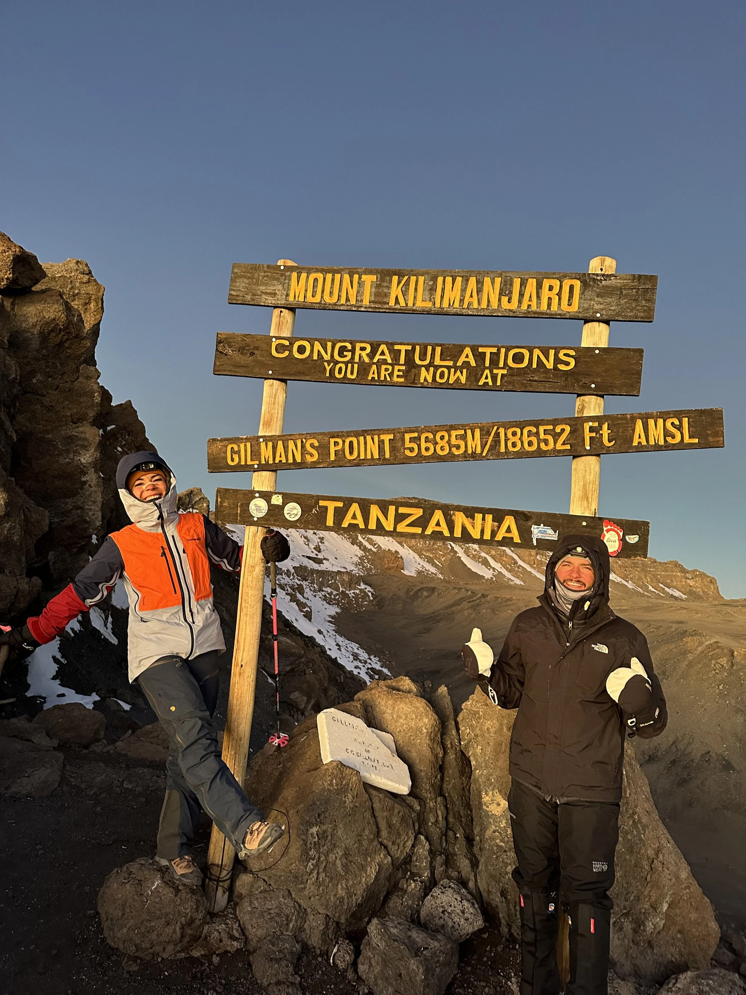 Two hikers at the summit of Mount Kilimanjaro next to a sign marking the peak at 5,685 meters (18,652 feet) above sea level in Tanzania, with snow patches and rocky terrain in the background.