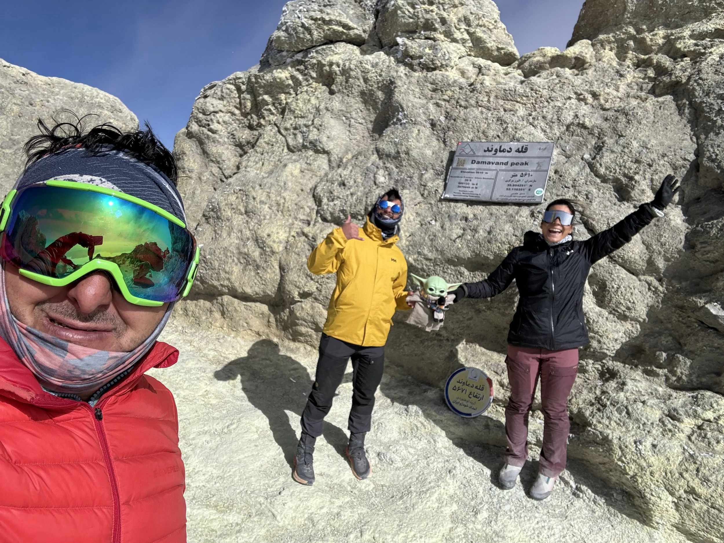 Three hikers at Damavand Peak, with a man in a red jacket and ski goggles taking a selfie, a person in a yellow jacket giving a thumbs-up, and a woman in a black jacket holding a small toy, standing in front of a sign and rocks.
