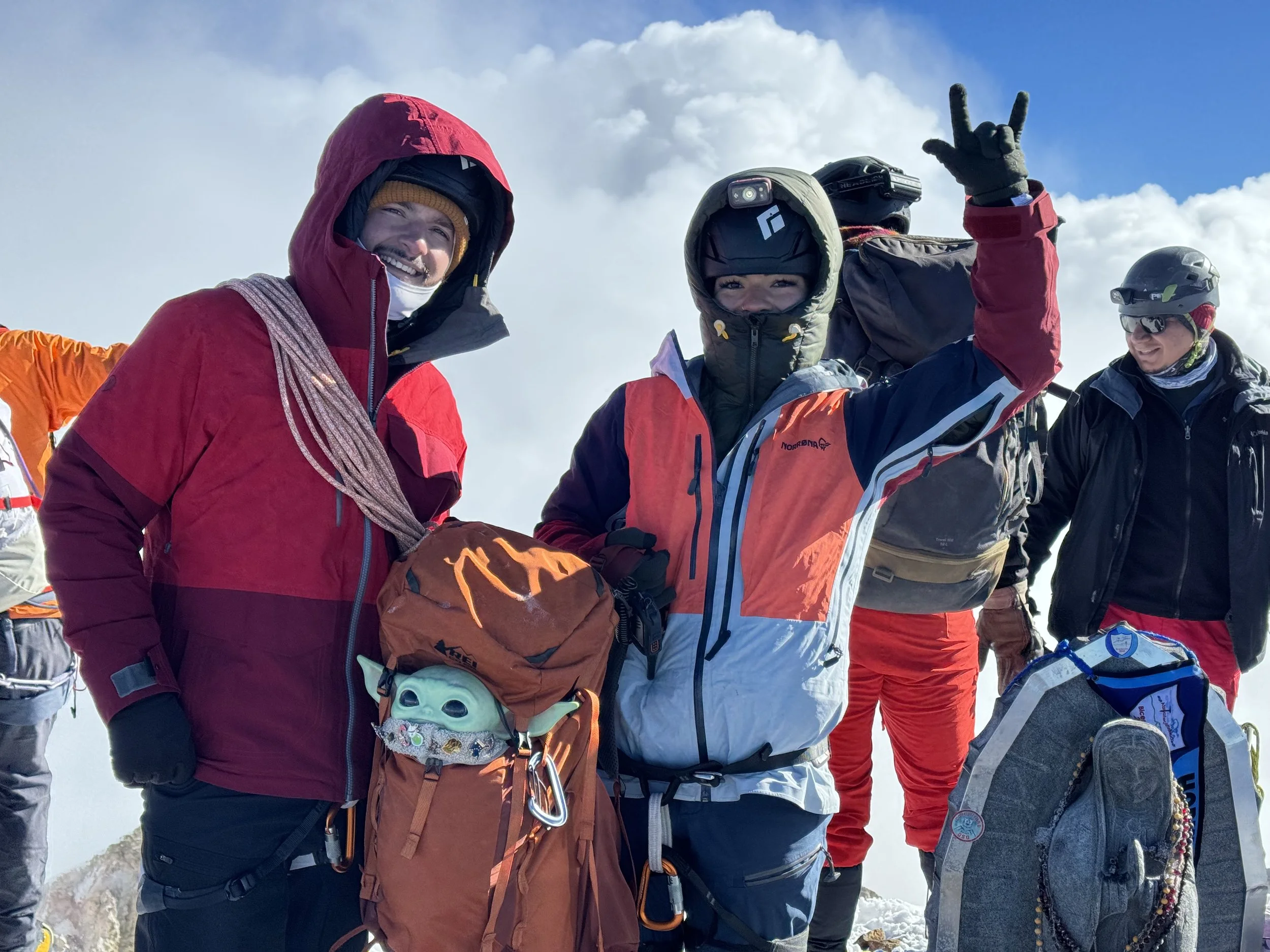 Group of mountain climbers dressed in colorful outdoor gear on snowy mountain summit, some smiling and making peace signs.