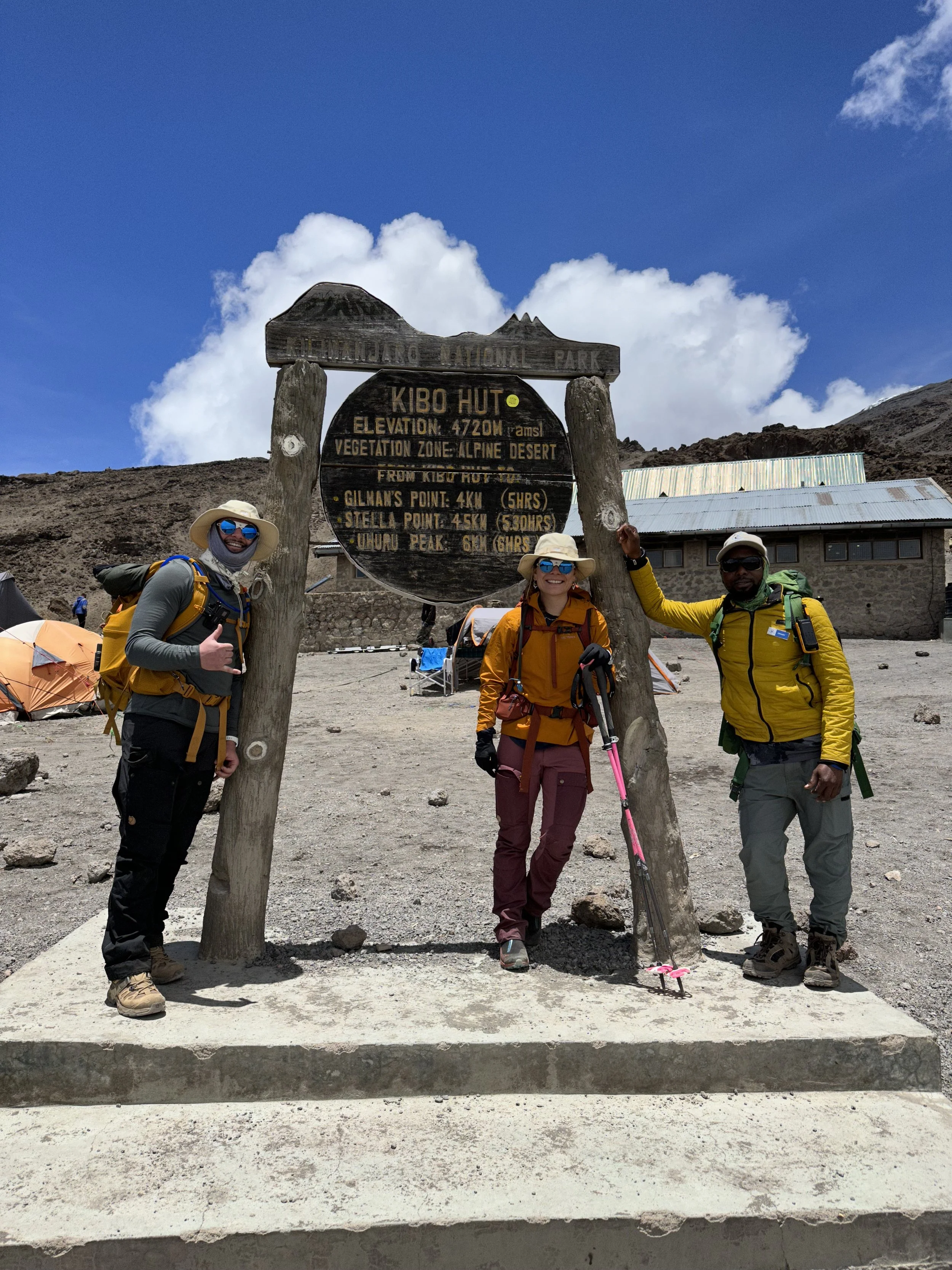 Three hikers in outdoor gear standing next to a wooden sign at Kibo Hut in Mount Kilimanjaro area. The sign indicates an elevation of 4720 meters and shows distances to various points, with a partly cloudy sky in the background.