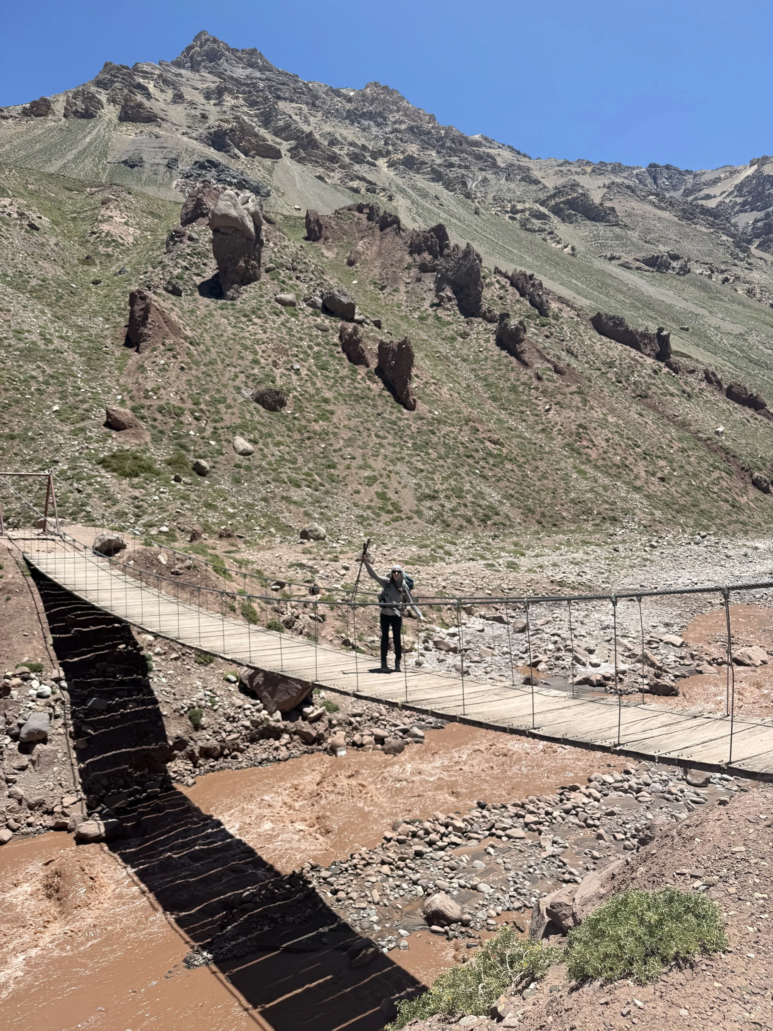 A person crossing a suspension bridge over a river in a mountainous, rocky landscape under a clear blue sky.