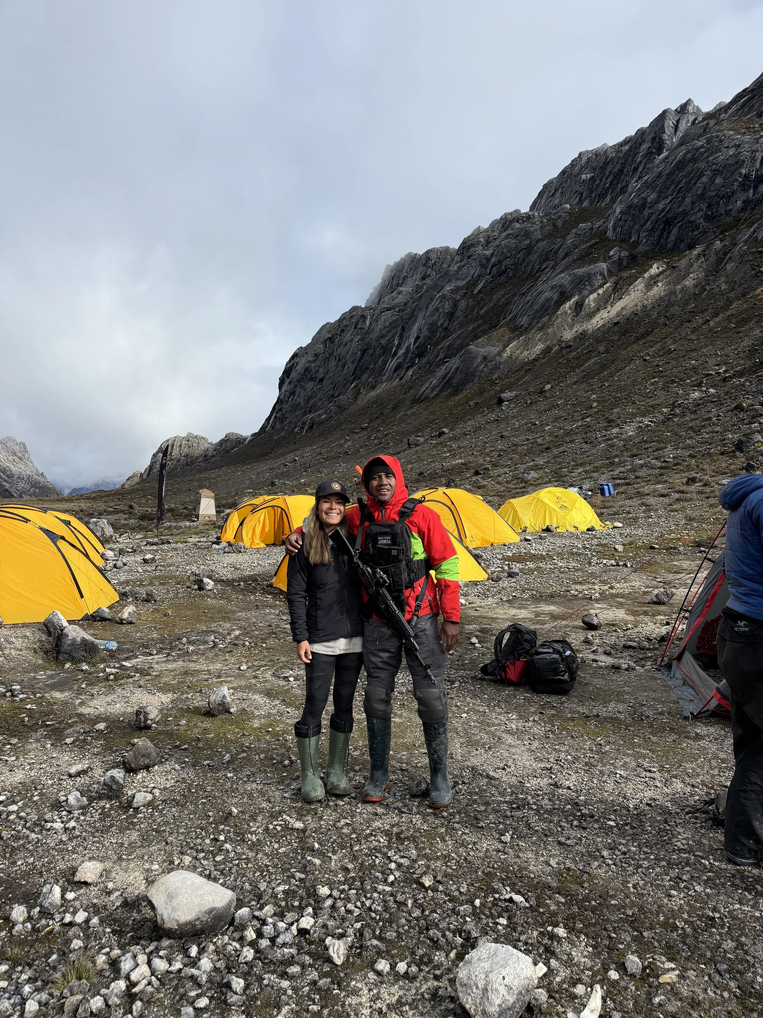 Two hikers, a woman and a man, are smiling and standing together on rocky ground in a mountain camp area with yellow tents, rocky mountain cliffs, and cloudy sky in the background.