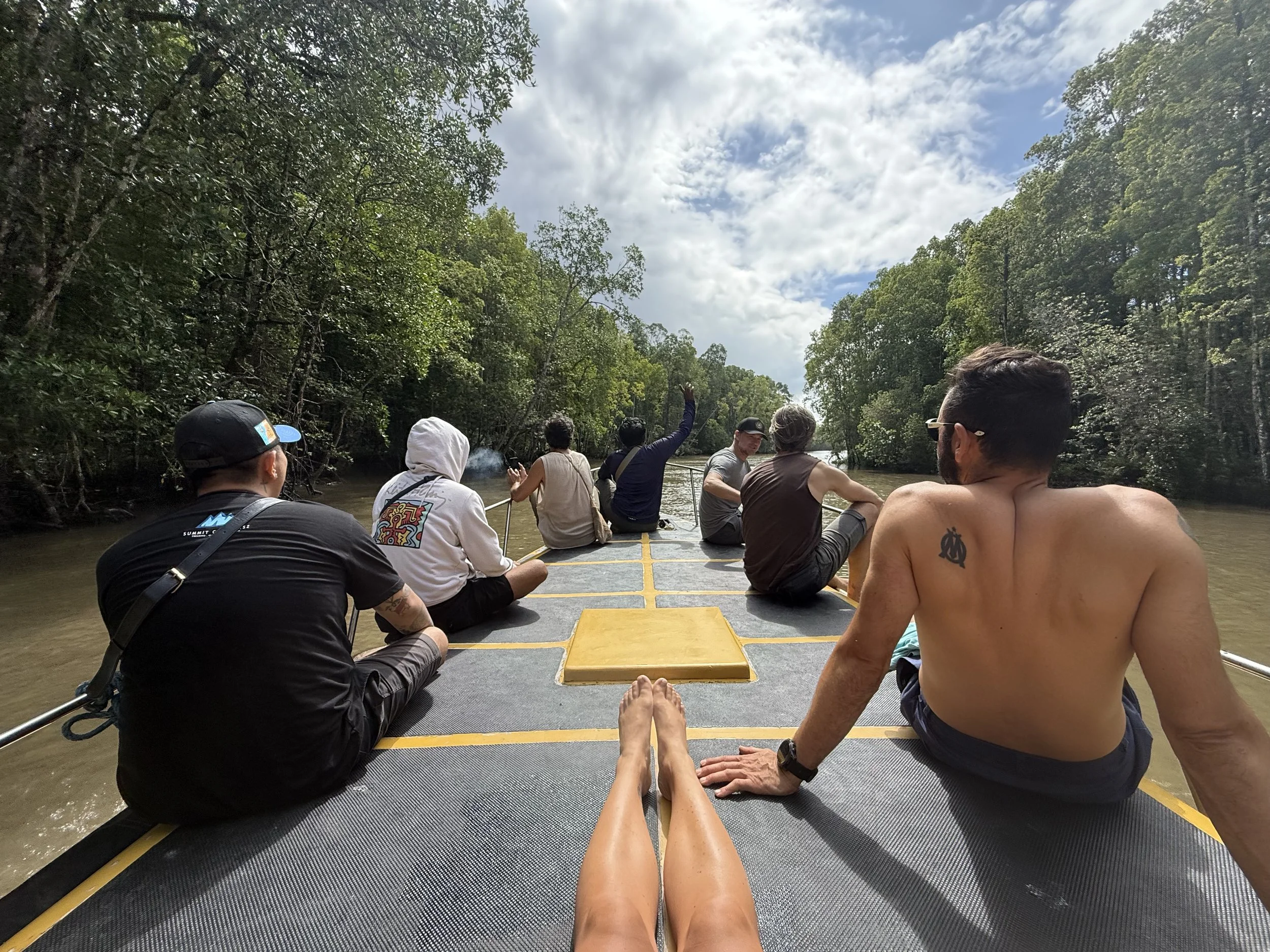 Group of people sitting on a boat floating down a river surrounded by lush green trees under a partly cloudy sky.