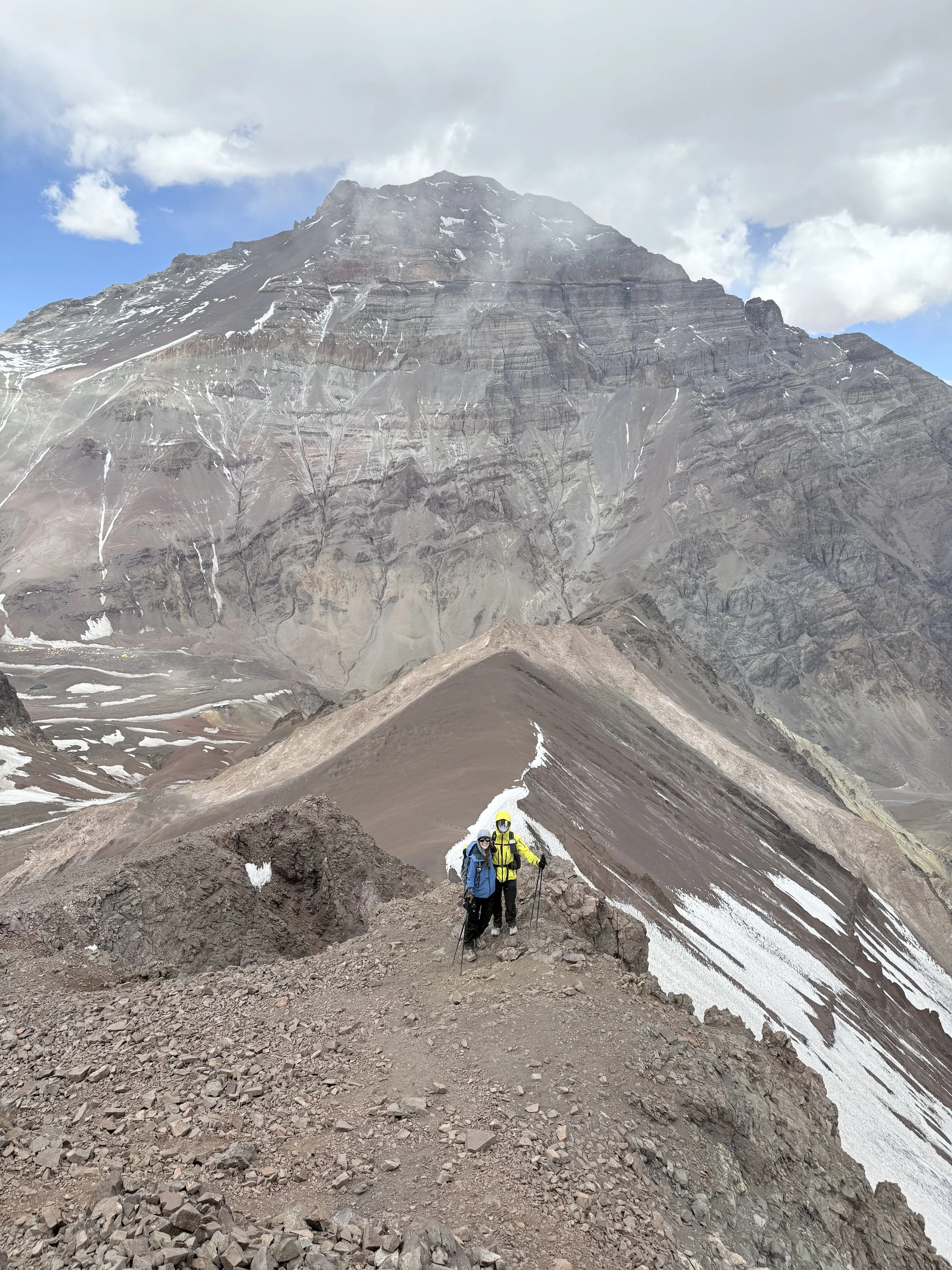 Two hikers in bright jackets and helmets standing on a mountain trail with a large rocky mountain in the background, partly covered with snow.