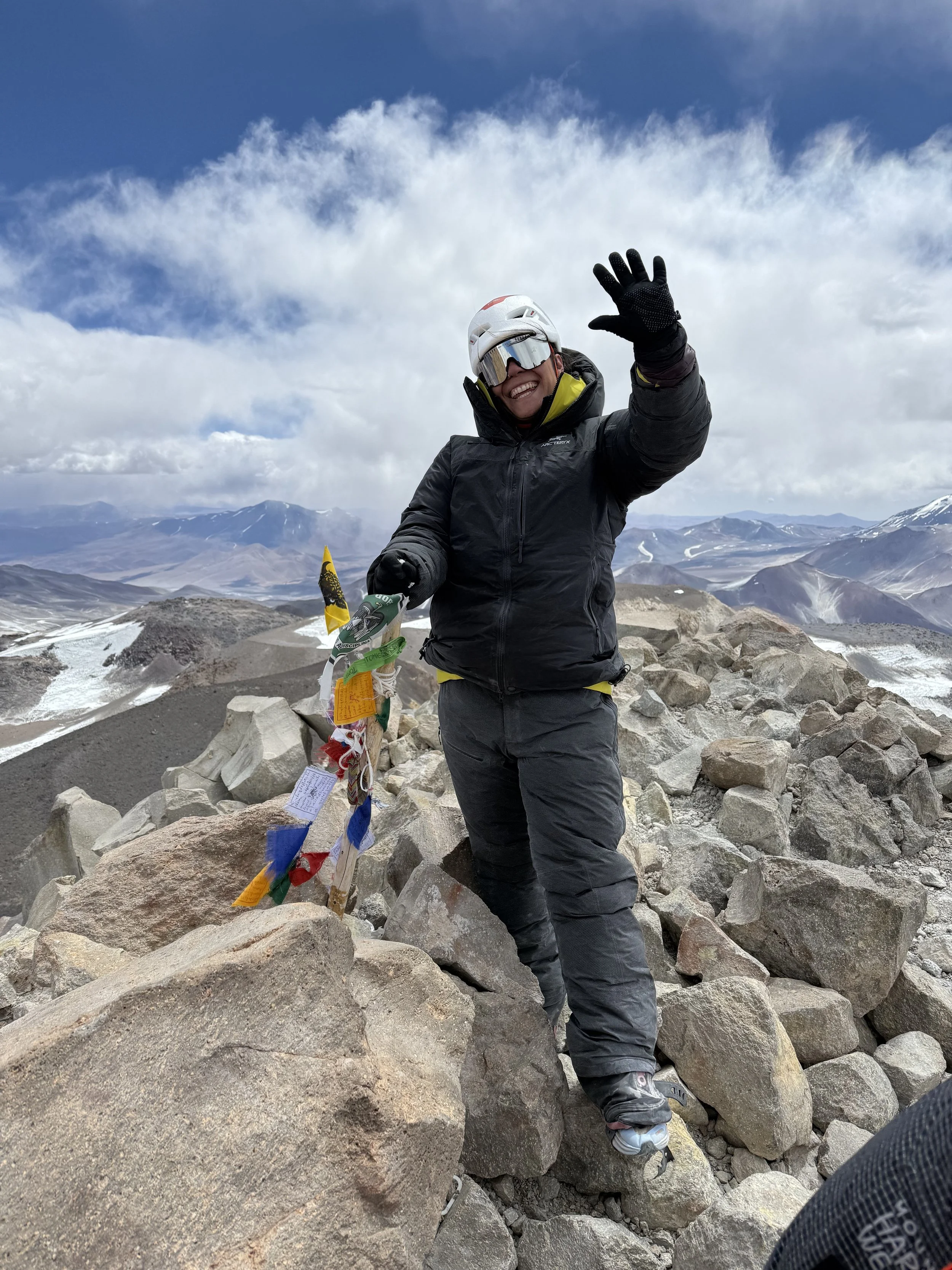 A person in mountaineering gear smiling and waving while standing on rocky terrain at a mountain summit, with snow-capped peaks and a partly cloudy sky in the background.