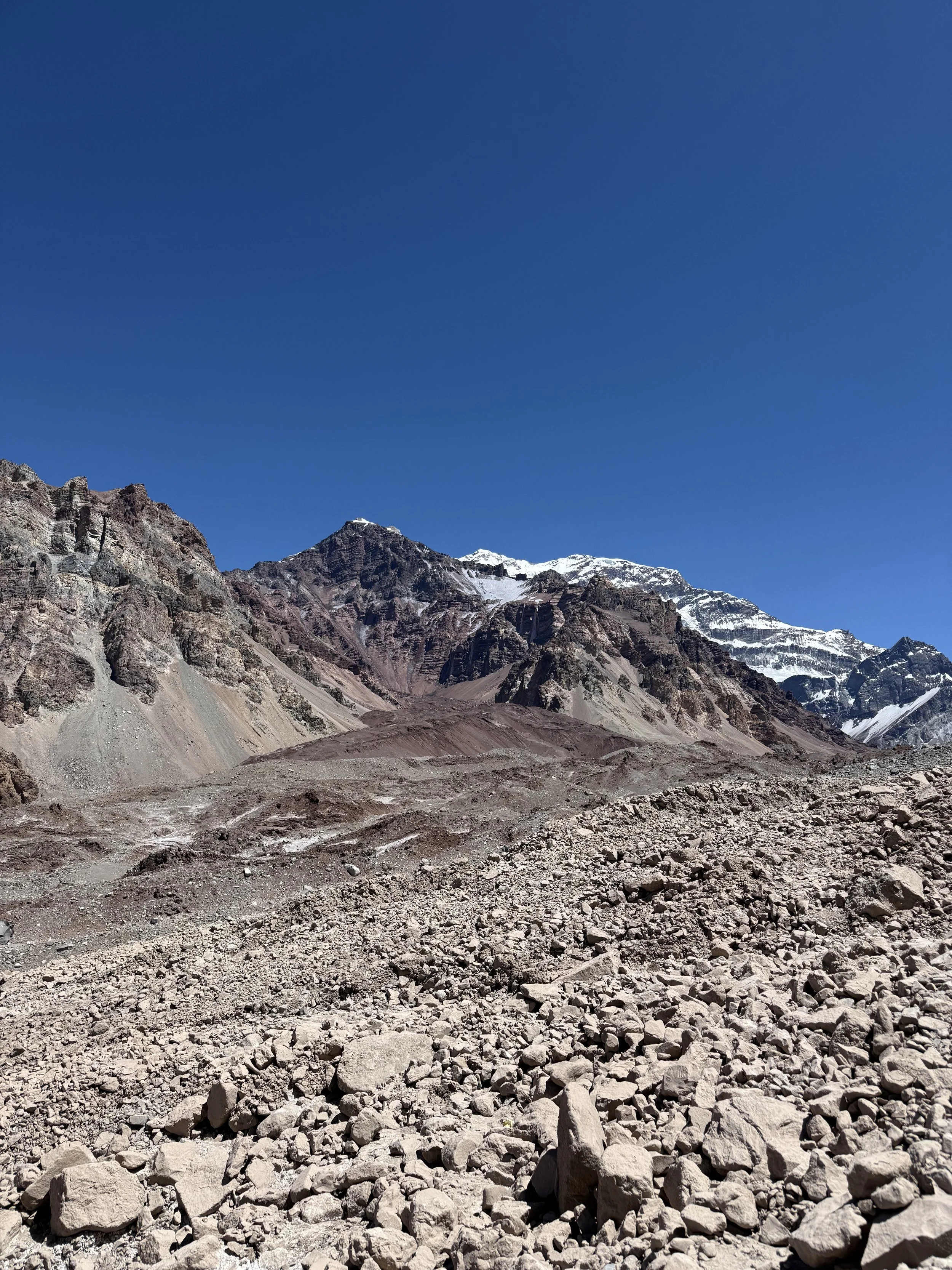 A rugged mountain landscape with rocky terrain in the foreground, steep mountain slopes in the middle, and snow-capped peaks under a clear blue sky in the background.