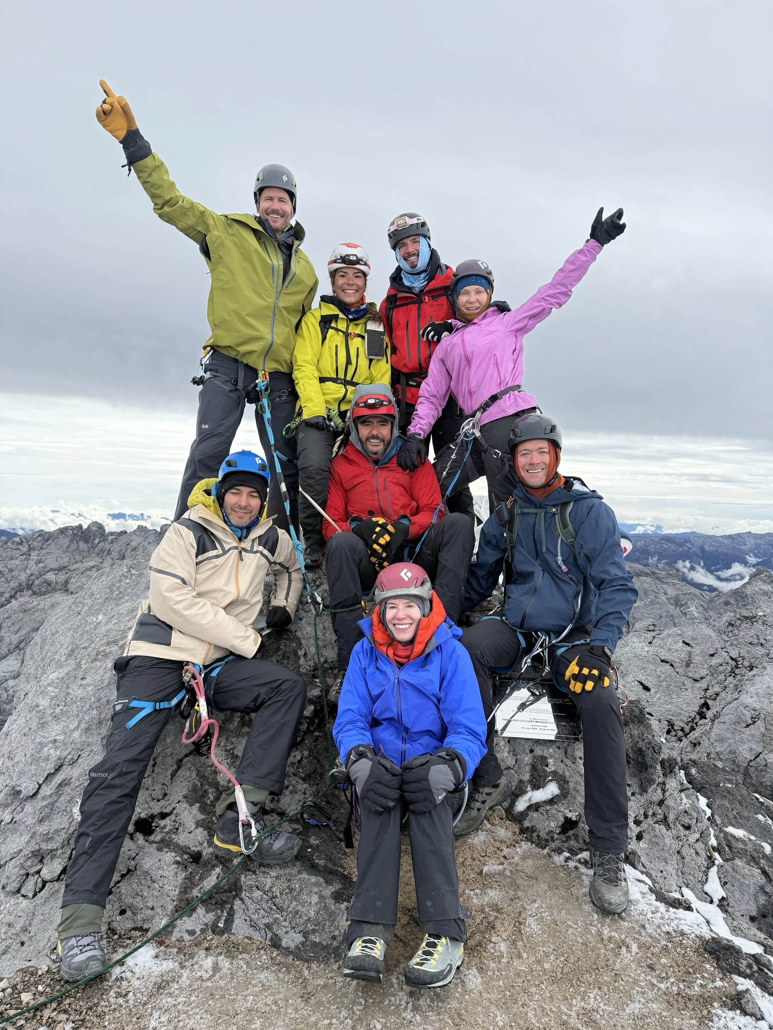 Group of nine climbers on a rocky mountain top, dressed in colorful outdoor gear and helmets, celebrating their ascent.