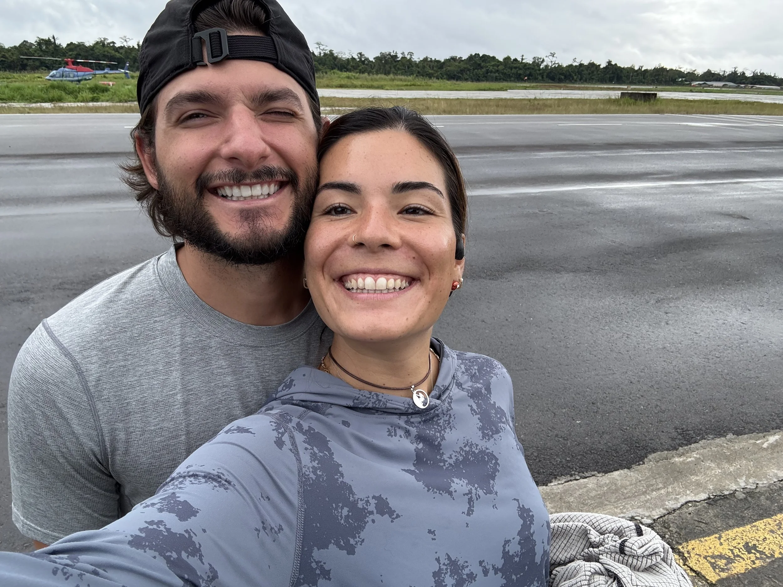A smiling man and woman taking a selfie outdoors on a rainy day near an airport runway with a helicopter and a small airplane in the background.
