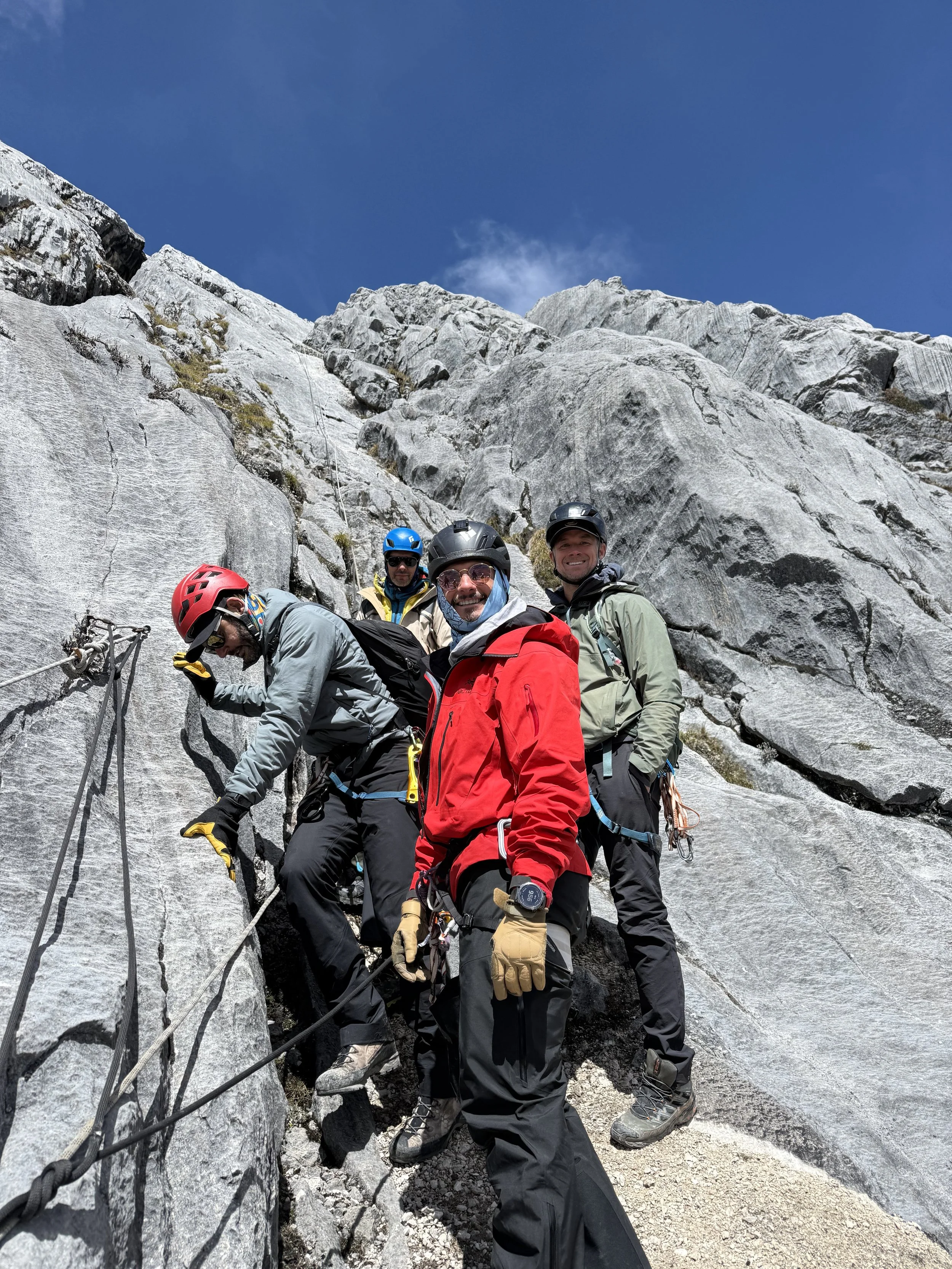 Four hikers wearing helmets and outdoor gear climbing a steep rocky mountain with safety ropes and harnesses.