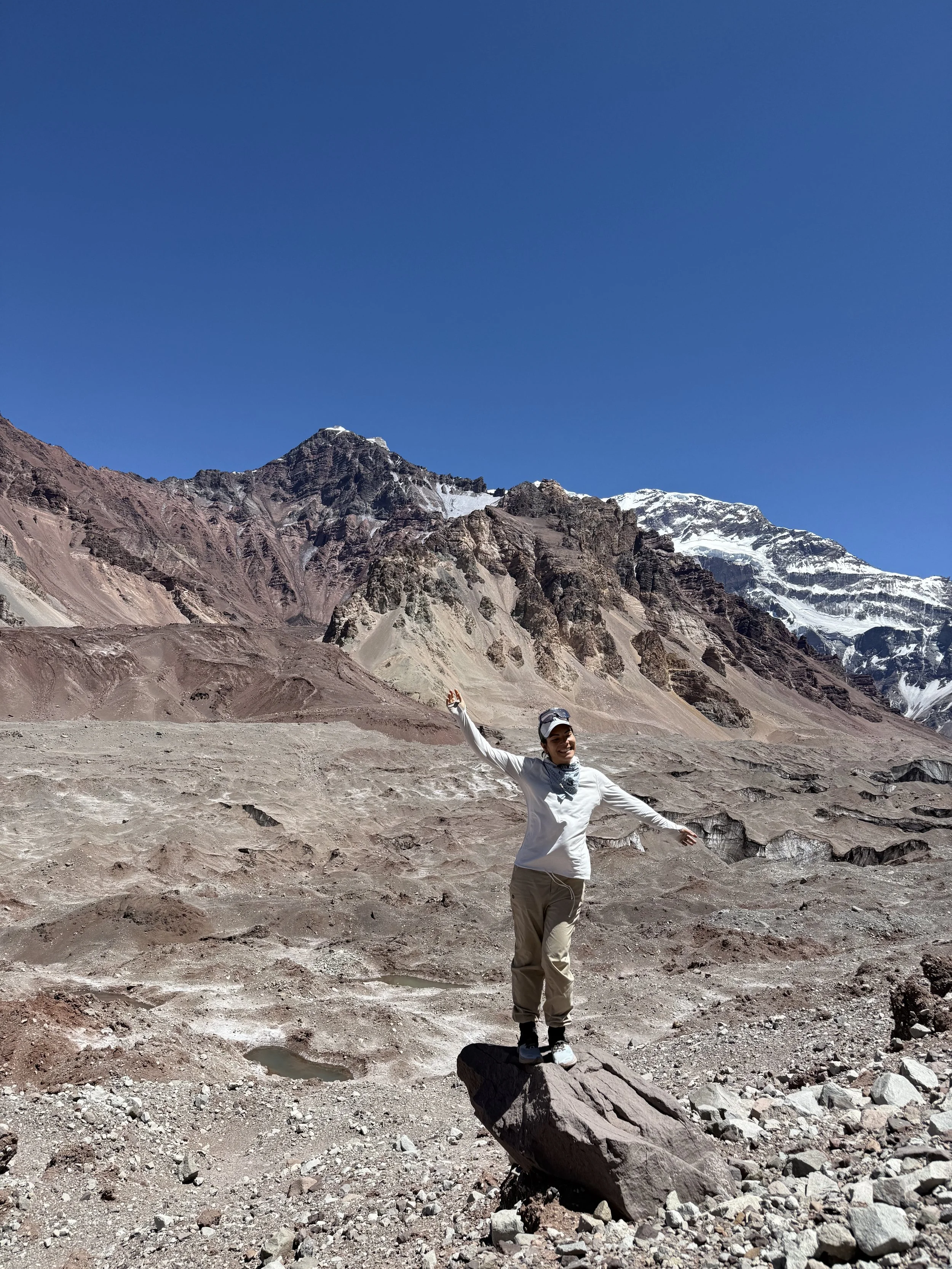 A woman standing on a large rock in a barren, rocky landscape with mountains and snow in the background, under a clear blue sky.