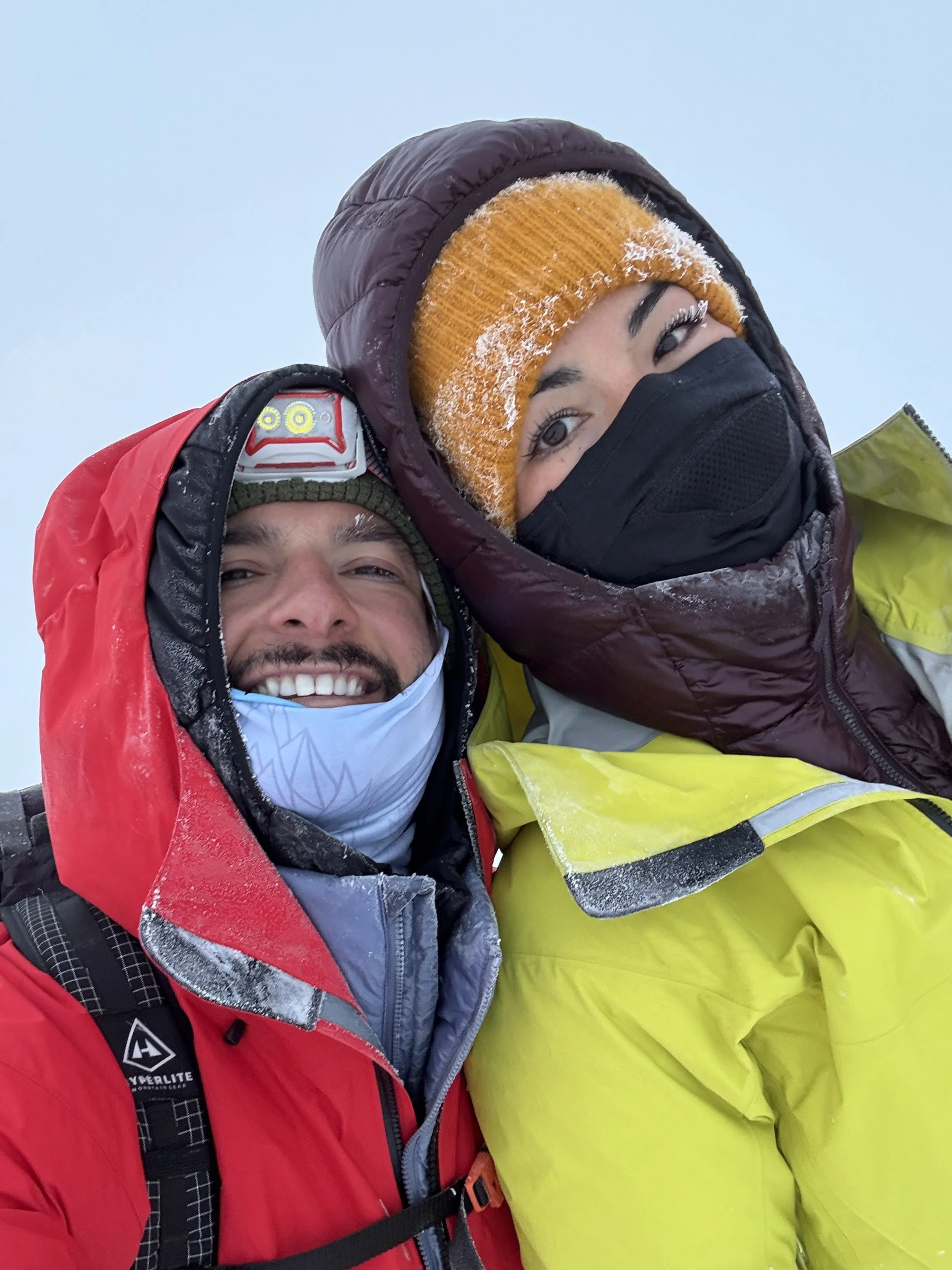 Two people dressed in winter clothing, smiling and taking a selfie in a snowy environment.