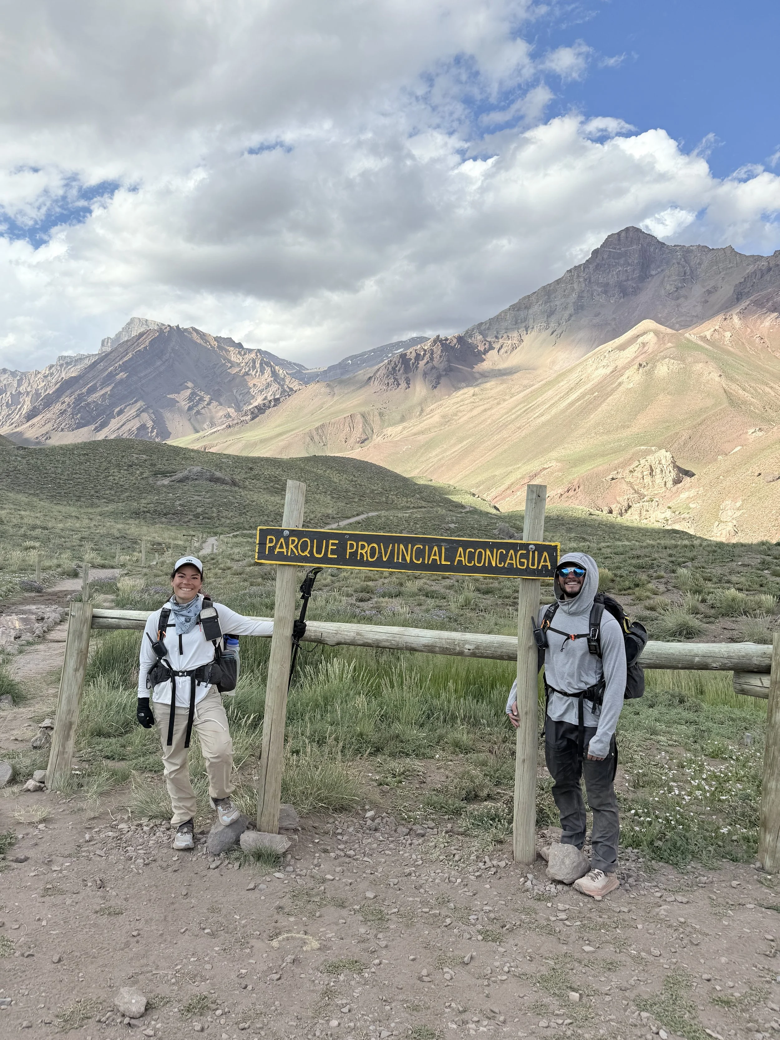 Two hikers standing near a sign for Parque Provincial Aconqugua, with mountains and a partly cloudy sky in the background.