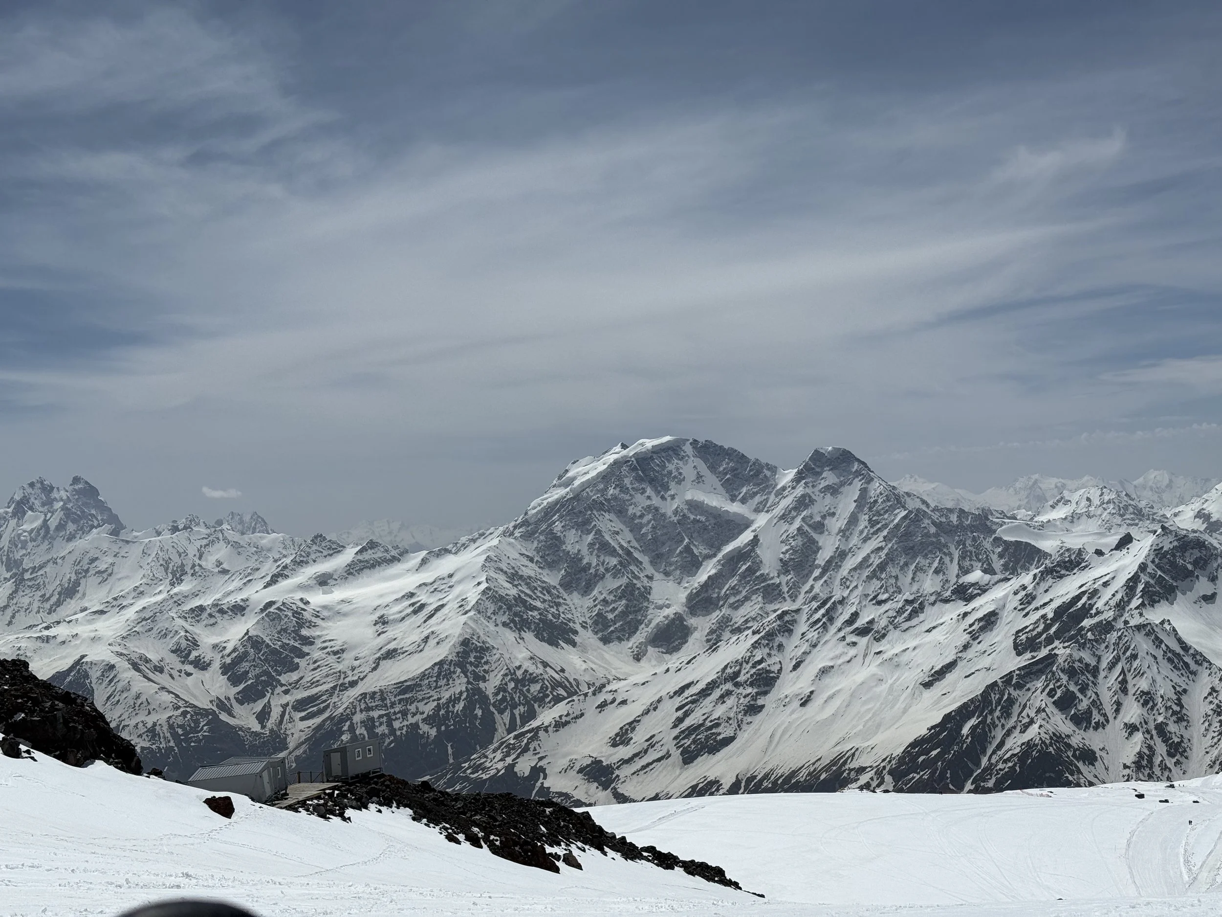 Snow-covered mountain range under a partly cloudy sky with a building and a snow-covered slope in the foreground.