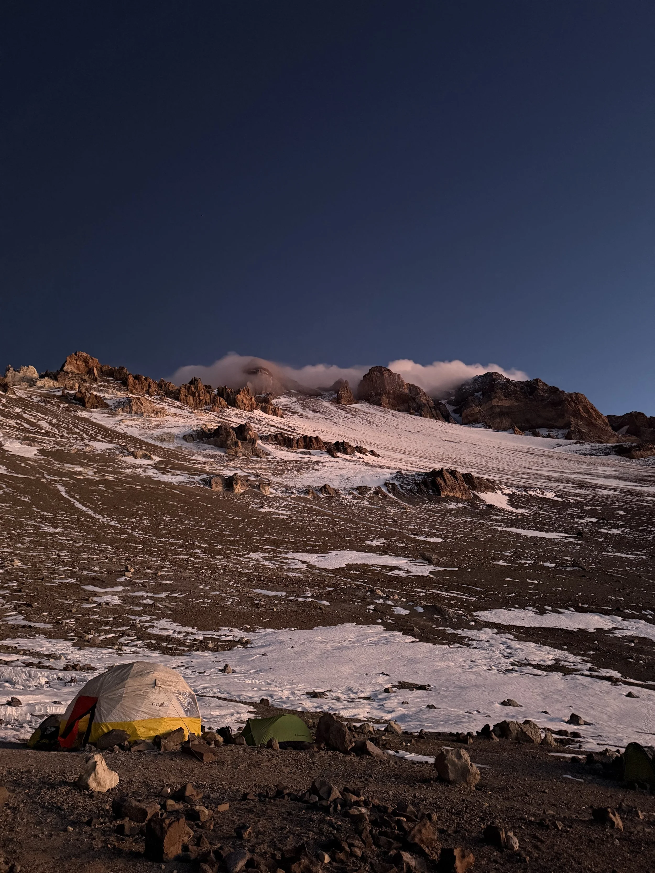 Tent camping on rocky terrain at the base of snow-covered mountains under a clear evening sky.