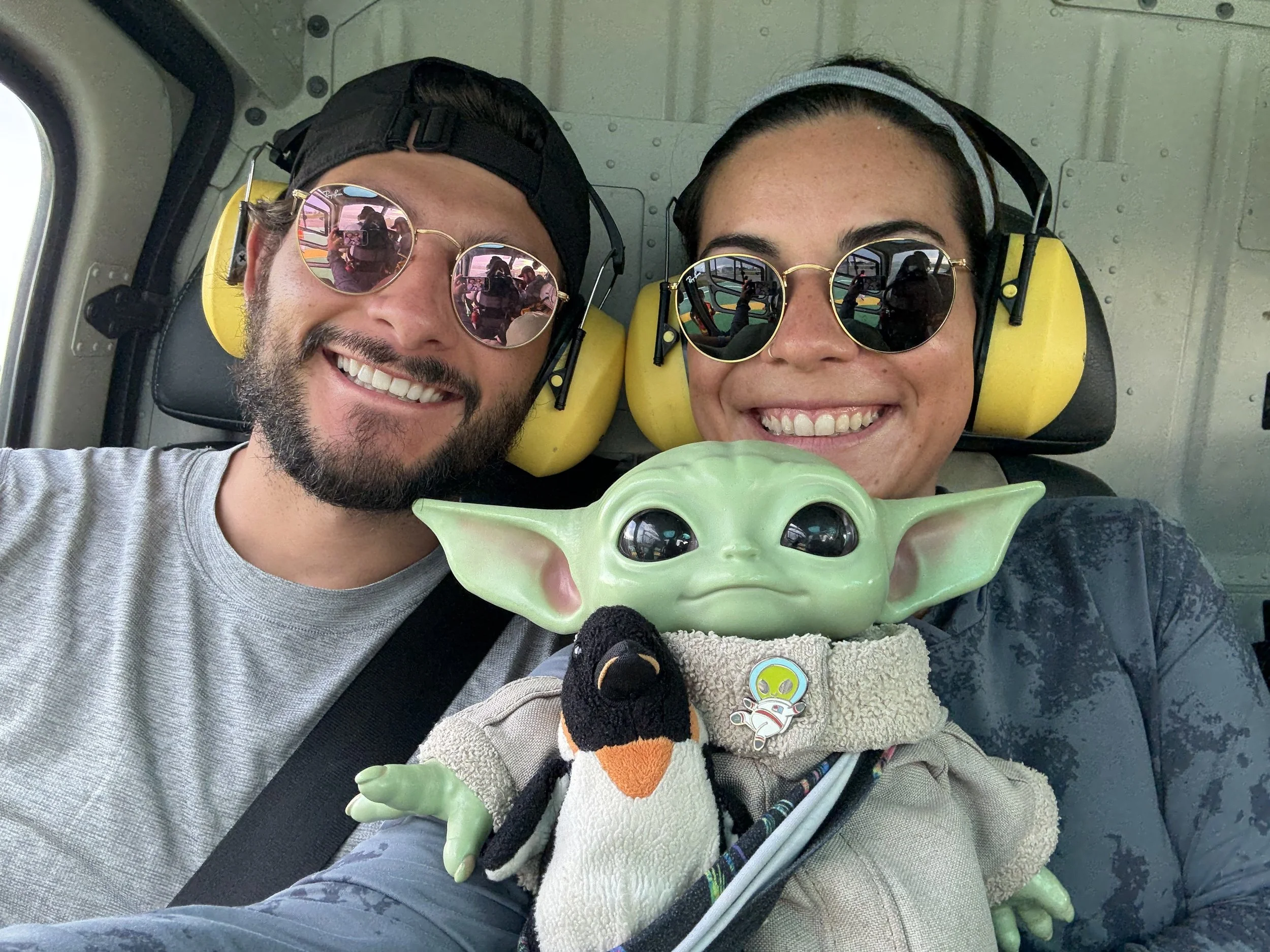 Two people are taking a selfie inside a helicopter, both wearing sunglasses and ear protection, with a Baby Yoda puppet and a plush penguin in the foreground.