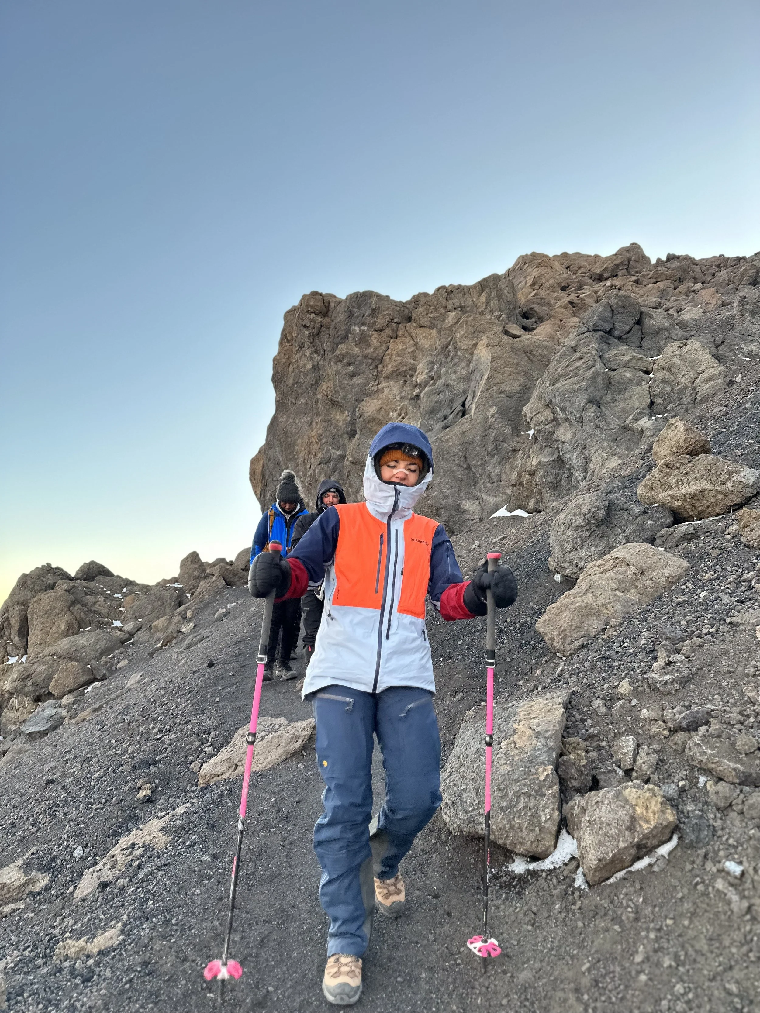 Three people in cold-weather hiking gear ascending a rocky mountain slope, with clear blue sky in the background.