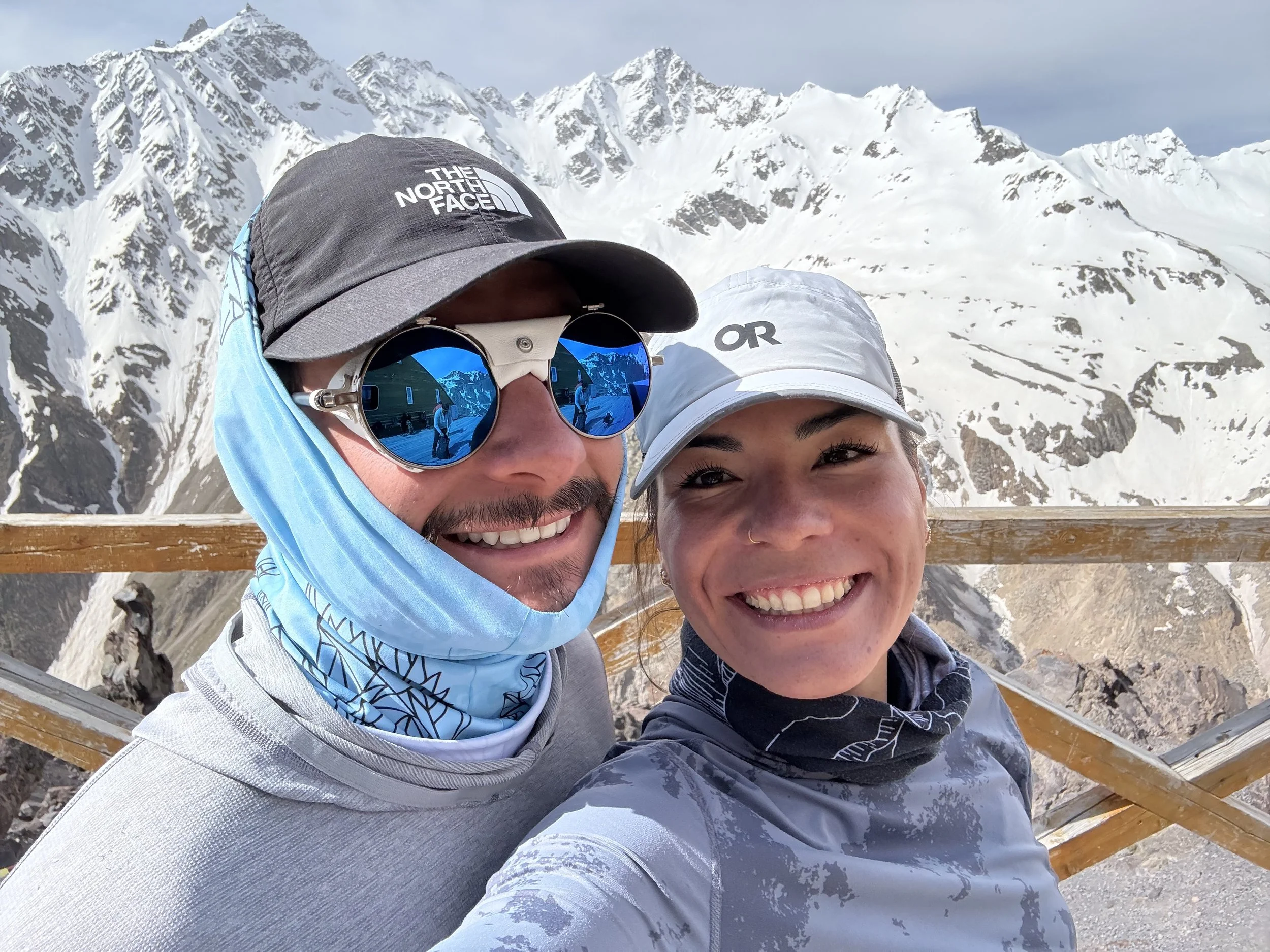 A smiling couple taking a selfie in a snowy mountain landscape, with mountains in the background and a wooden railing in the foreground.