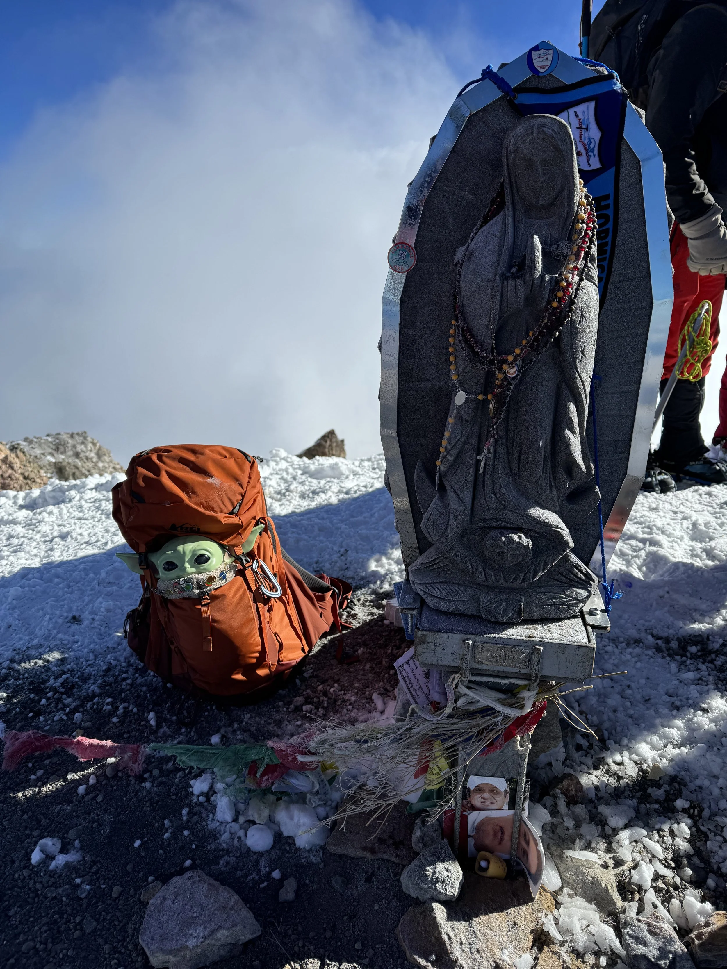 A memorial with a small green alien figure in an orange backpack, a small photo of a smiling person, and prayer beads, set on snow and rocks at the summit of a mountain.