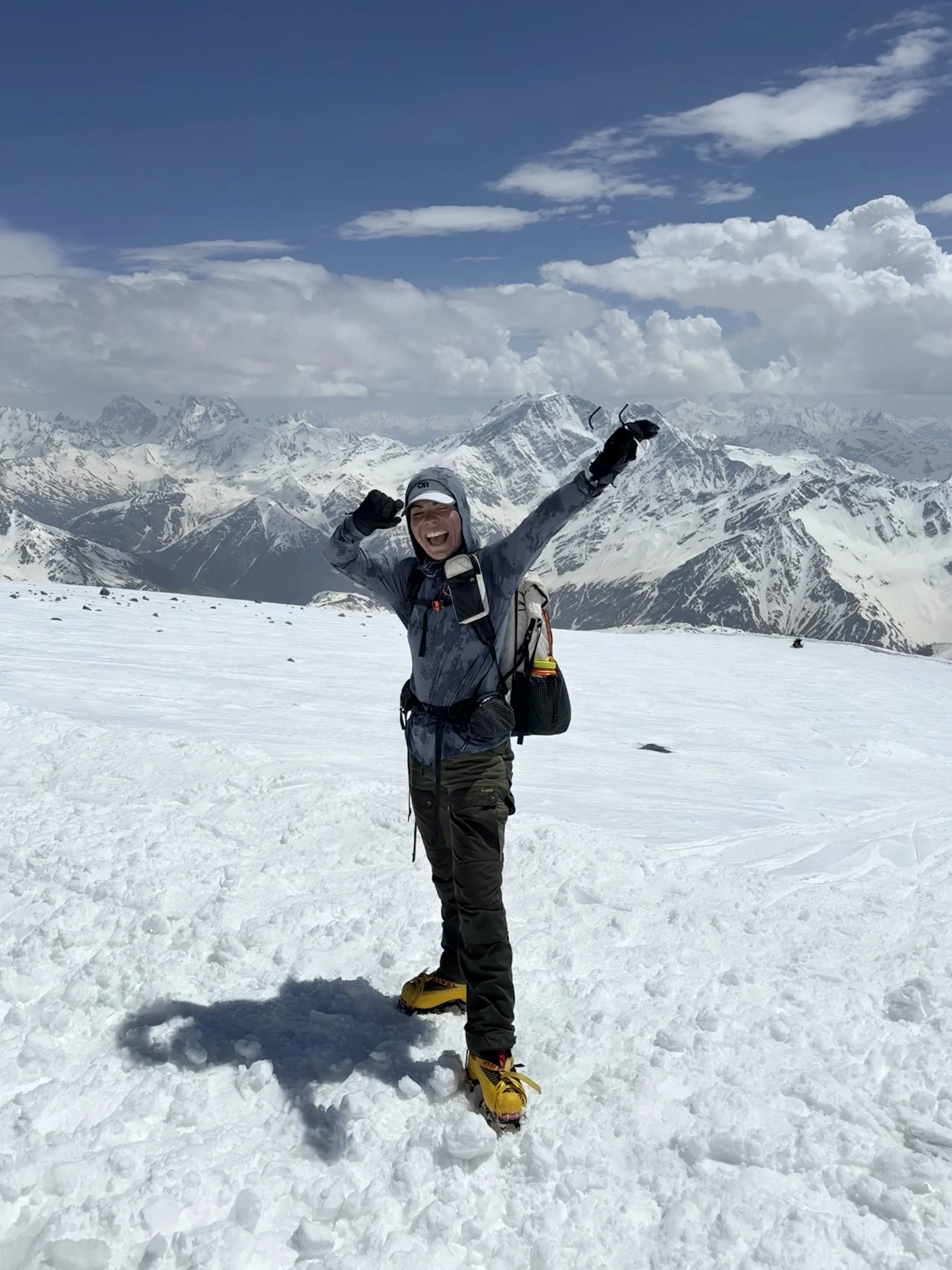 Person in winter gear standing on snow-covered mountain, smiling and cheering, with snow-capped mountains and blue sky with clouds in the background.