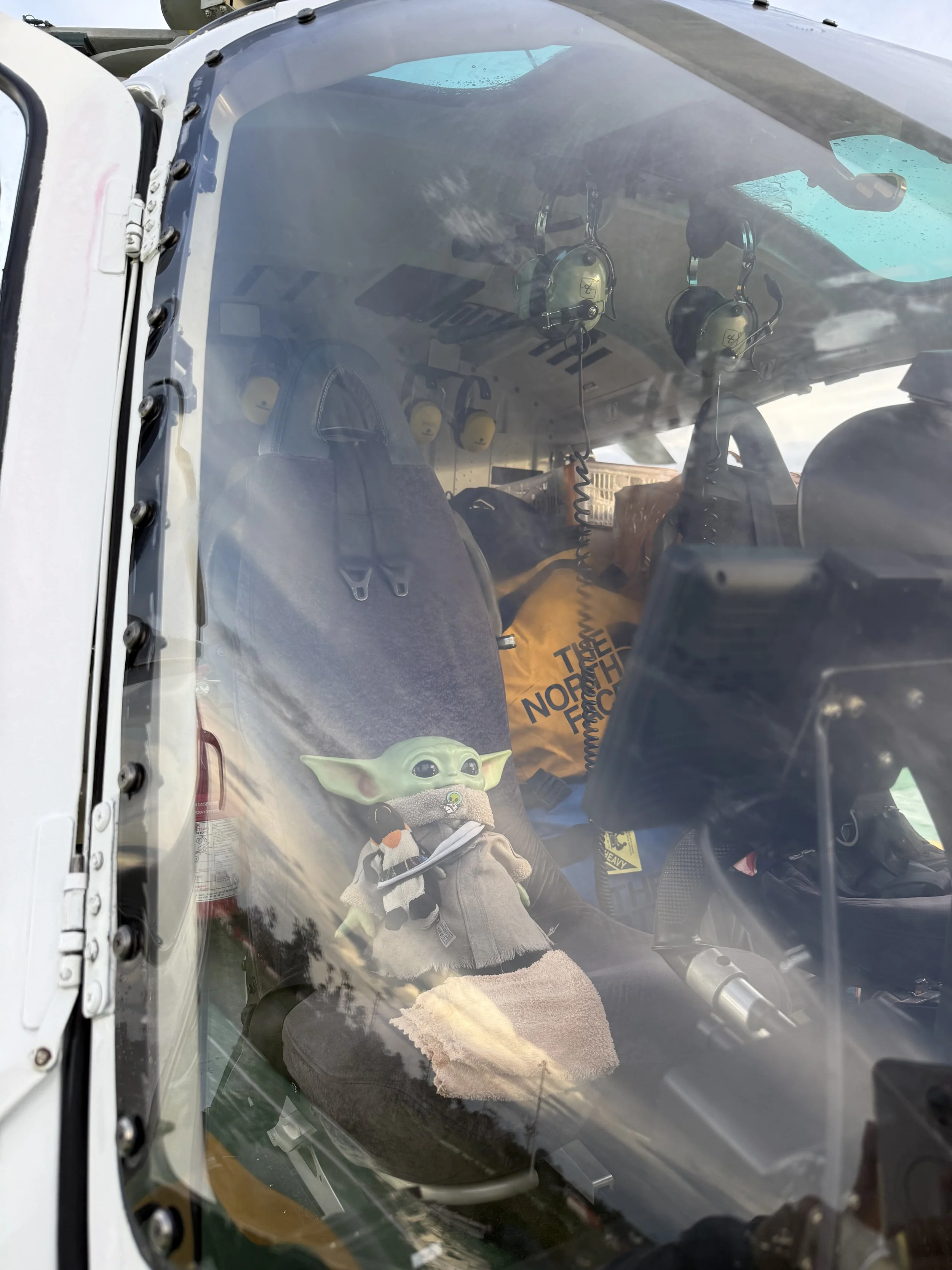 View through the cockpit window of a helicopter, showing the seats, multiple headsets hanging, a plush Baby Yoda toy dressed in grey, and a yellow North Face bag.