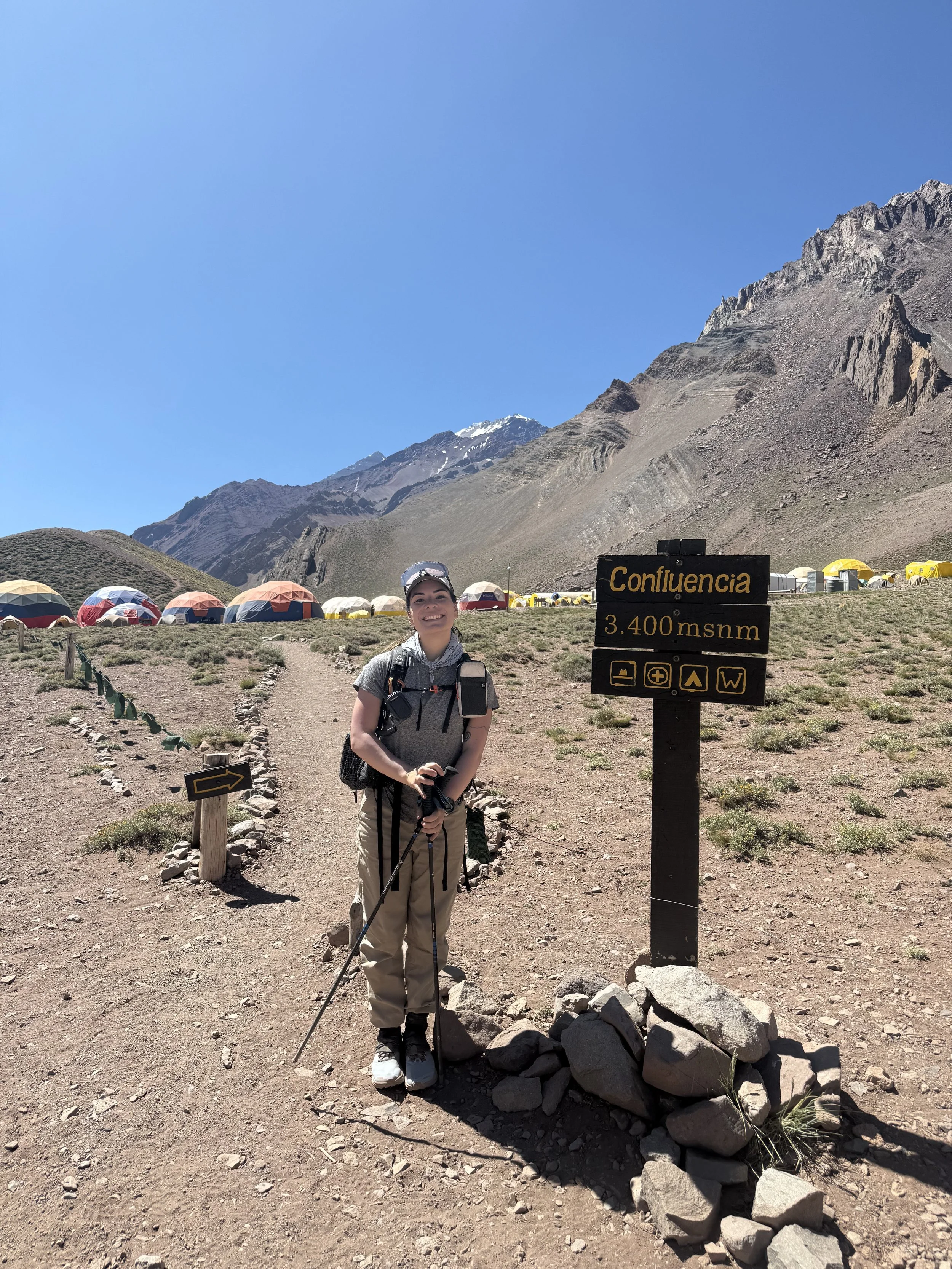 A woman standing on a mountain trail at Confluencia, 3,400 meters above sea level, with tents and rugged mountains in the background.
