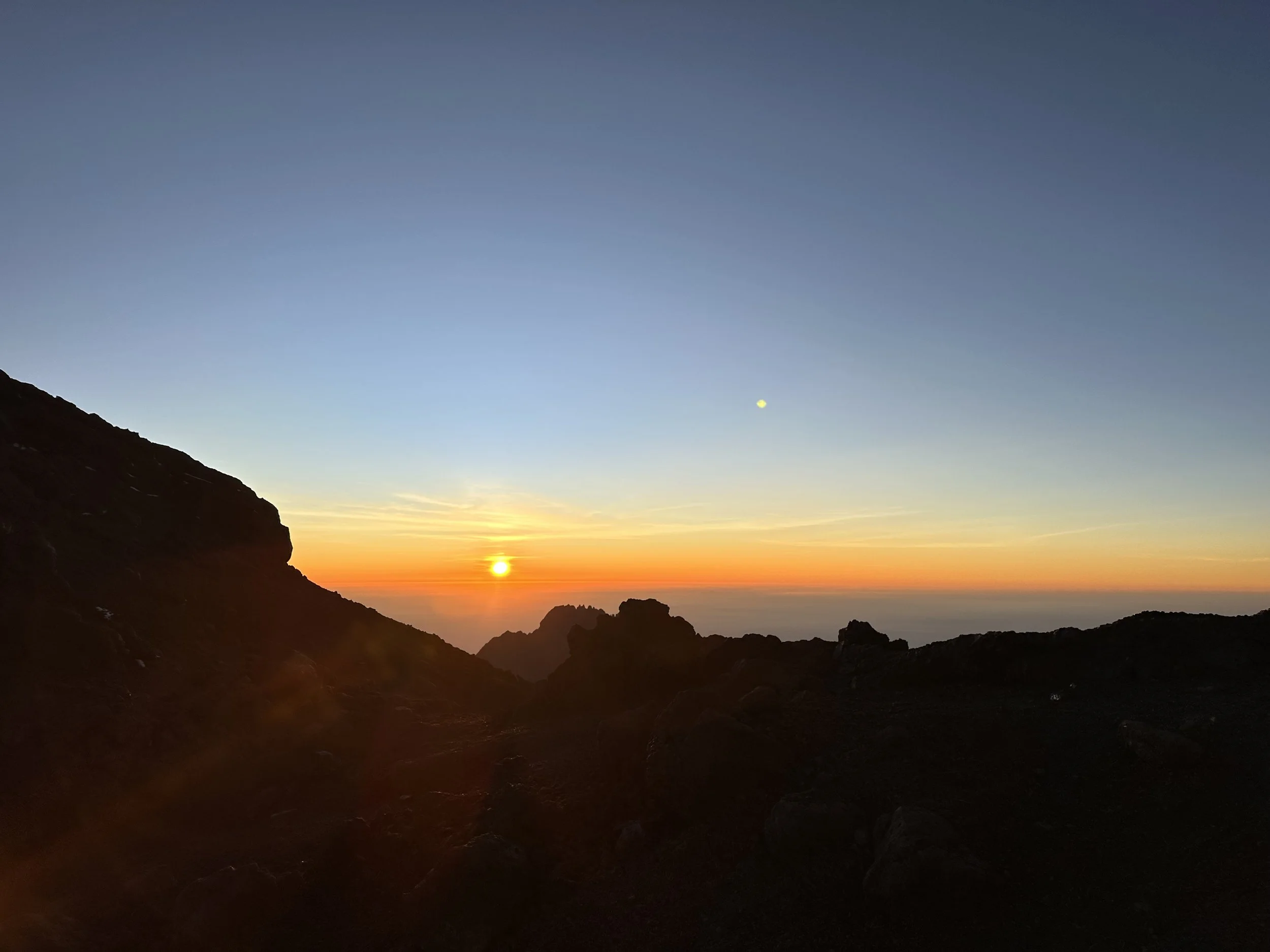 Sunset over a mountain landscape with a clear sky, silhouettes of rugged terrain, and the moon visible in the sky.