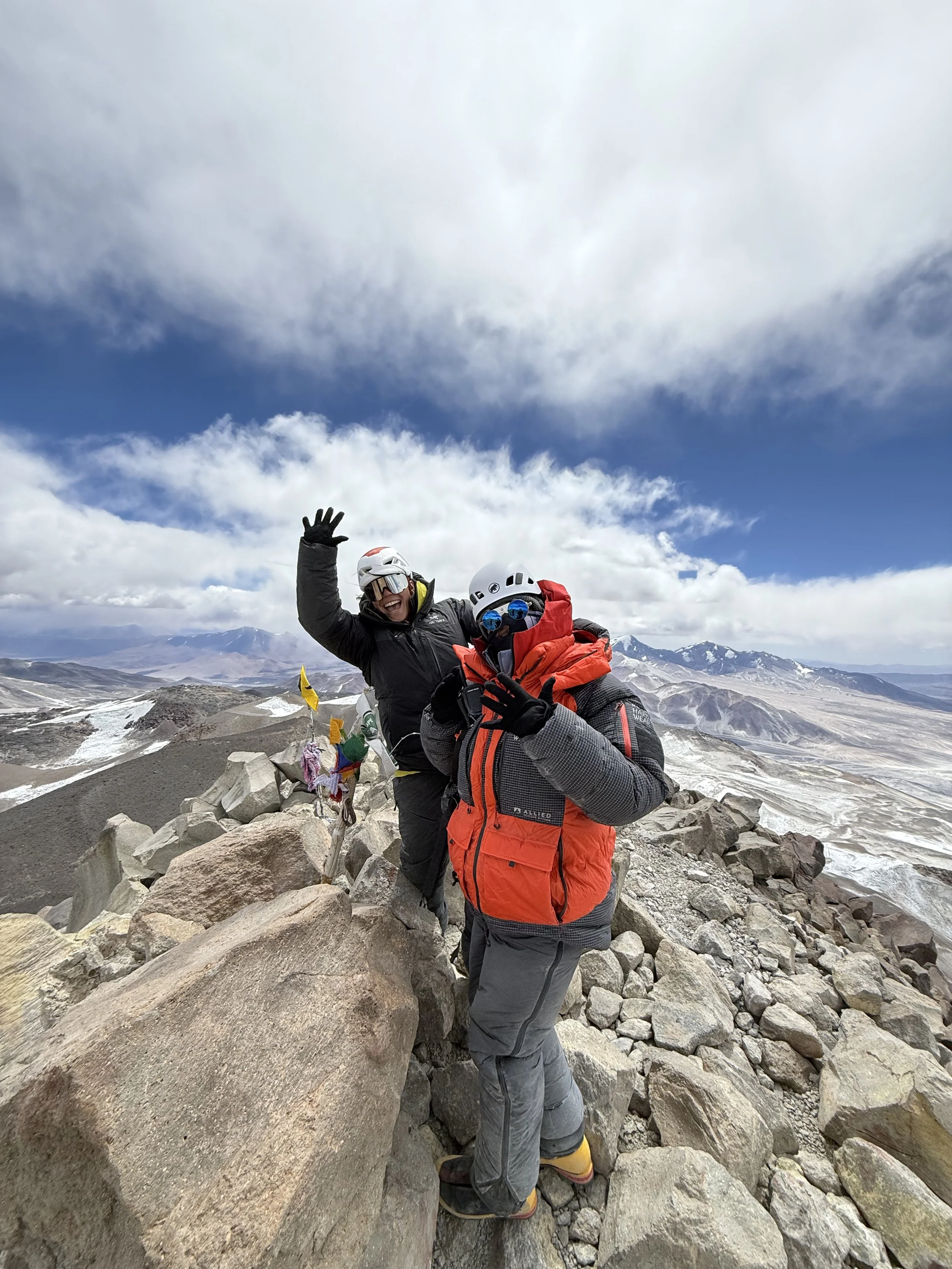 Two hikers wearing helmets and winter gear standing on rocky mountain terrain, smiling and waving at the camera with cloudy sky and distant mountain landscape in the background.