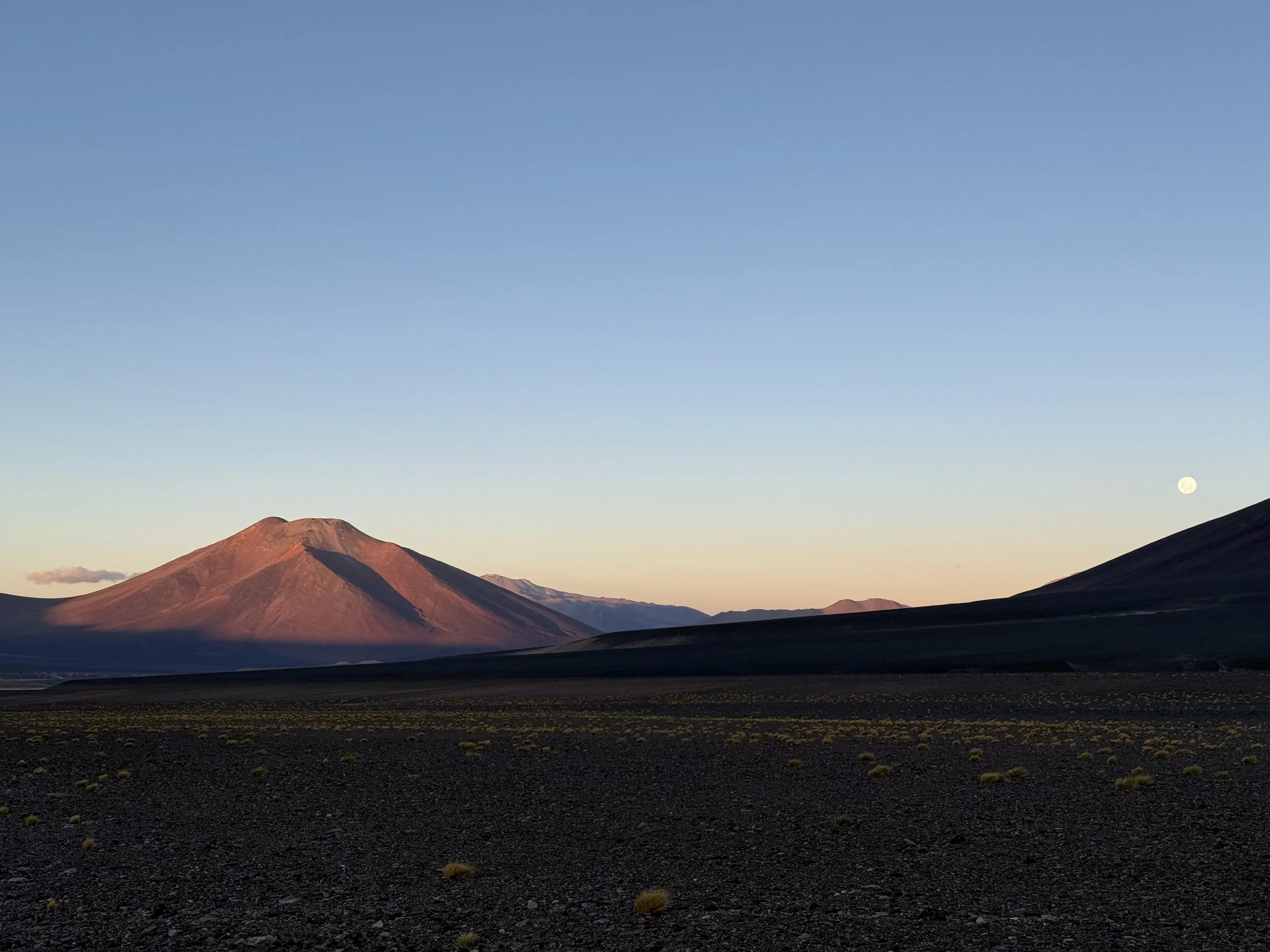 A desert landscape with dark volcanic terrain, a mountain with a reddish hue, a pale blue sky, and a visible moon in the distance.