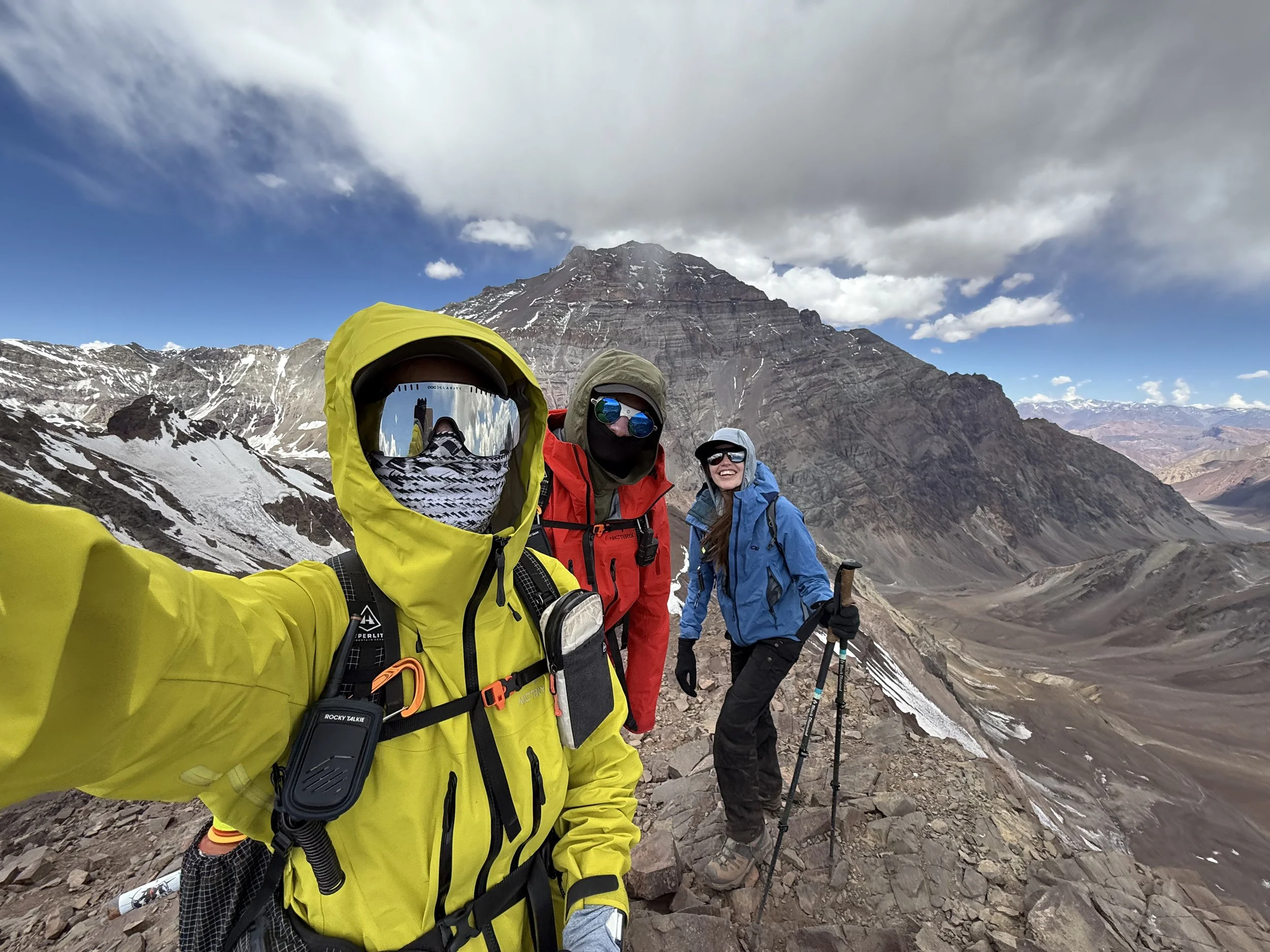 Three hikers wearing colorful jackets and protective gear taking a selfie on a mountain trail with snow-capped peaks and cloudy sky in the background.
