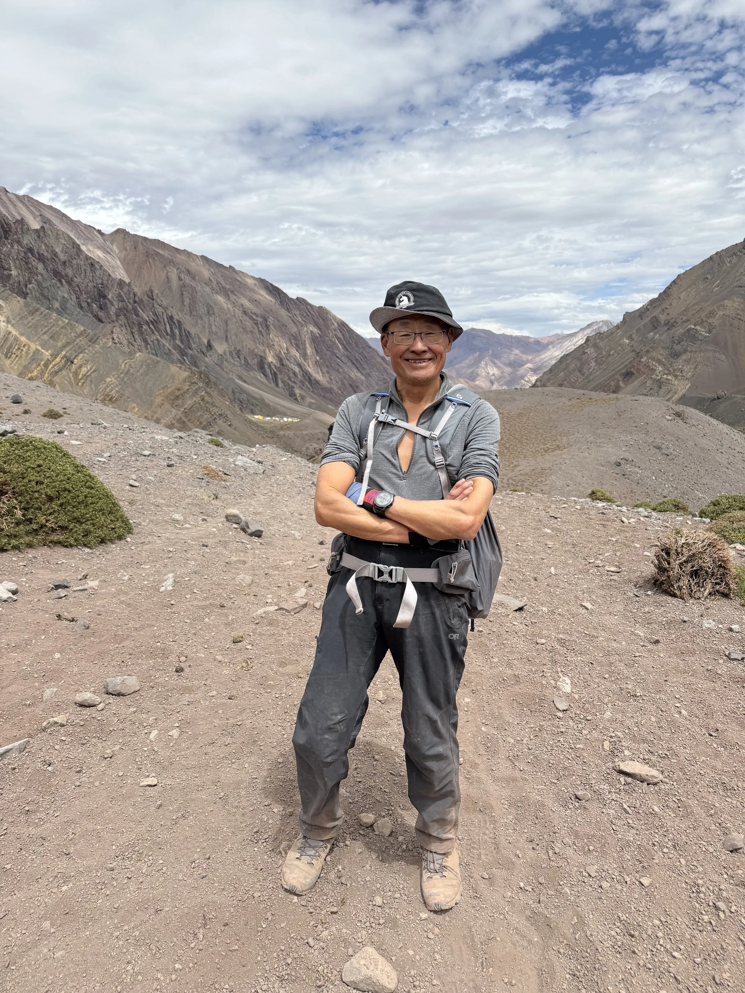 A smiling man in outdoor hiking gear standing on a dirt trail in a mountainous desert landscape on a cloudy day.