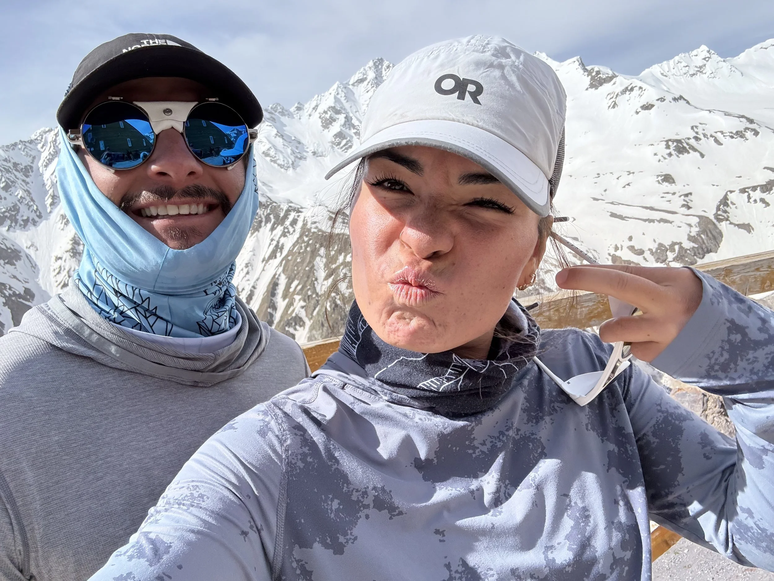 Two smiling friends taking a selfie with snow-covered mountains in the background, one woman wearing a white cap and gray jacket, the man wearing reflective sunglasses and a black cap.