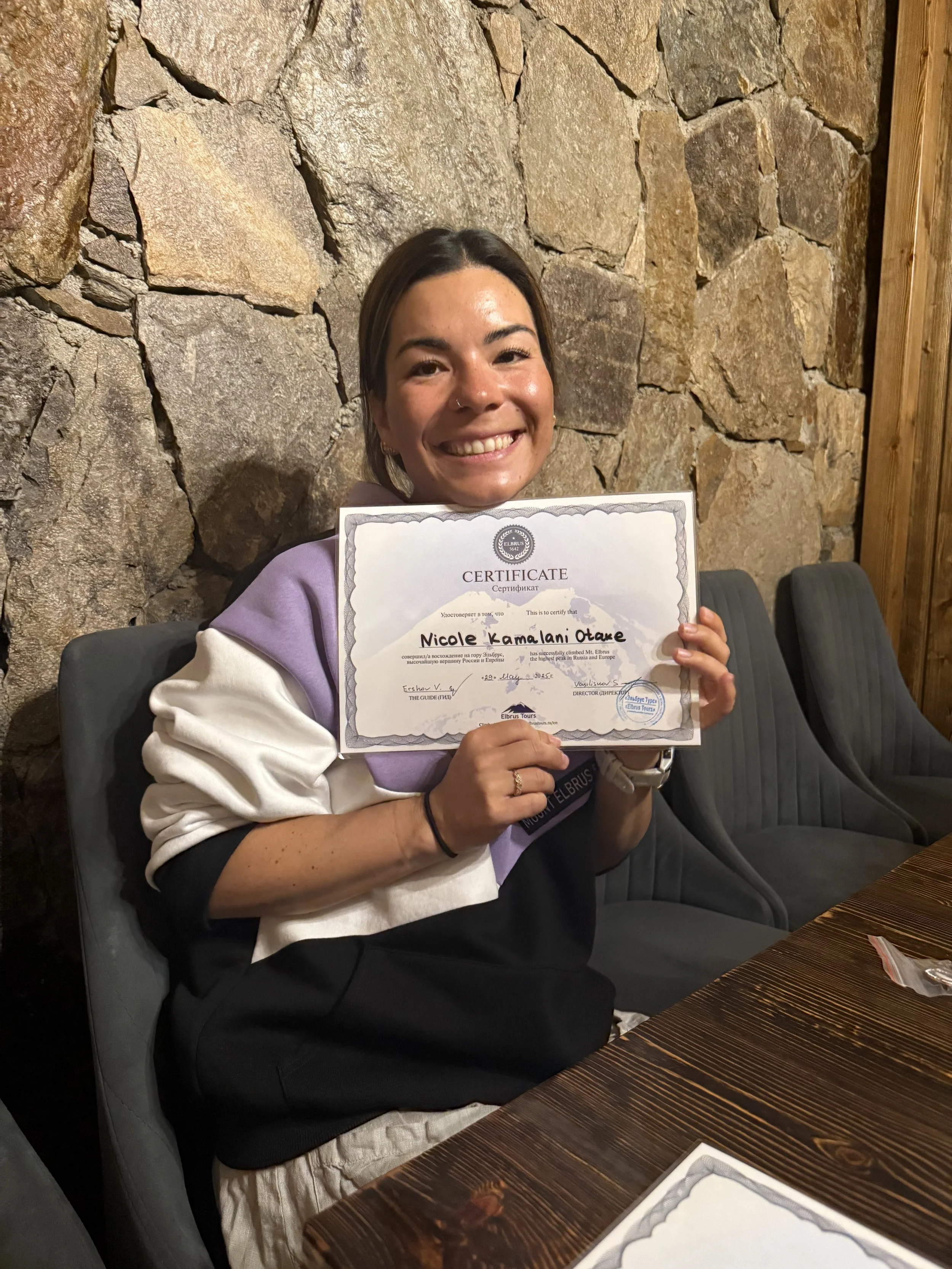 A young woman with dark hair and a nose ring smiling and holding a certificate at a table with a stone wall background.