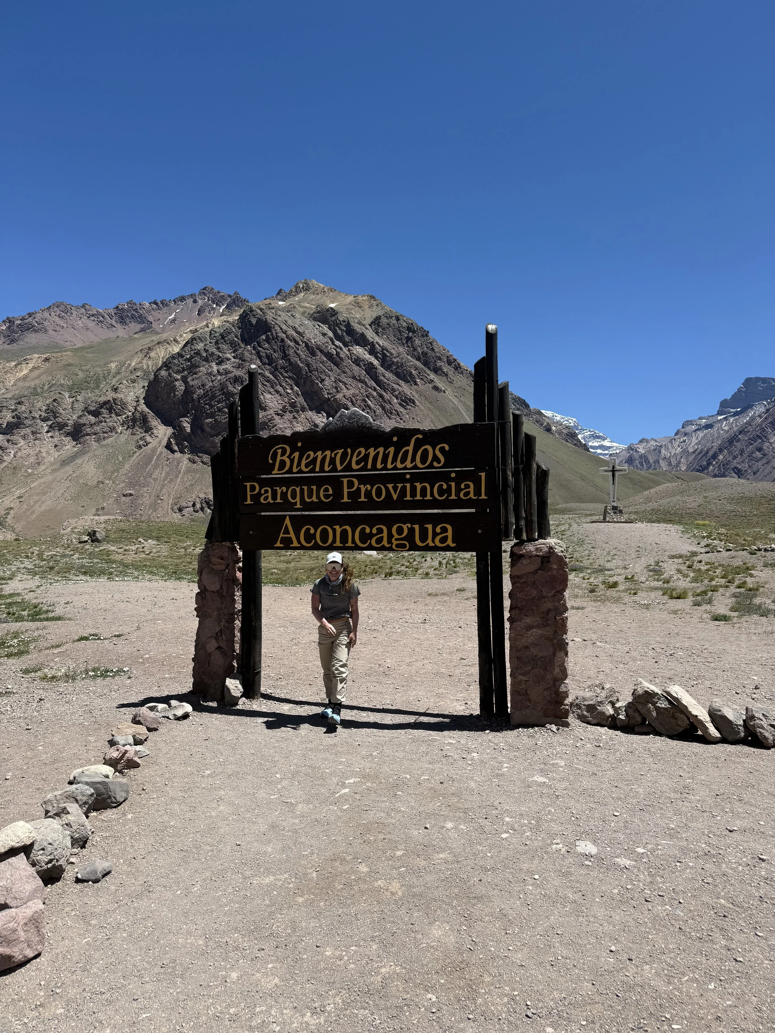 A woman standing under a wooden sign that reads 'Bienvenidos Parque Provincial Aconcagua' in a mountainous landscape with clear blue sky.