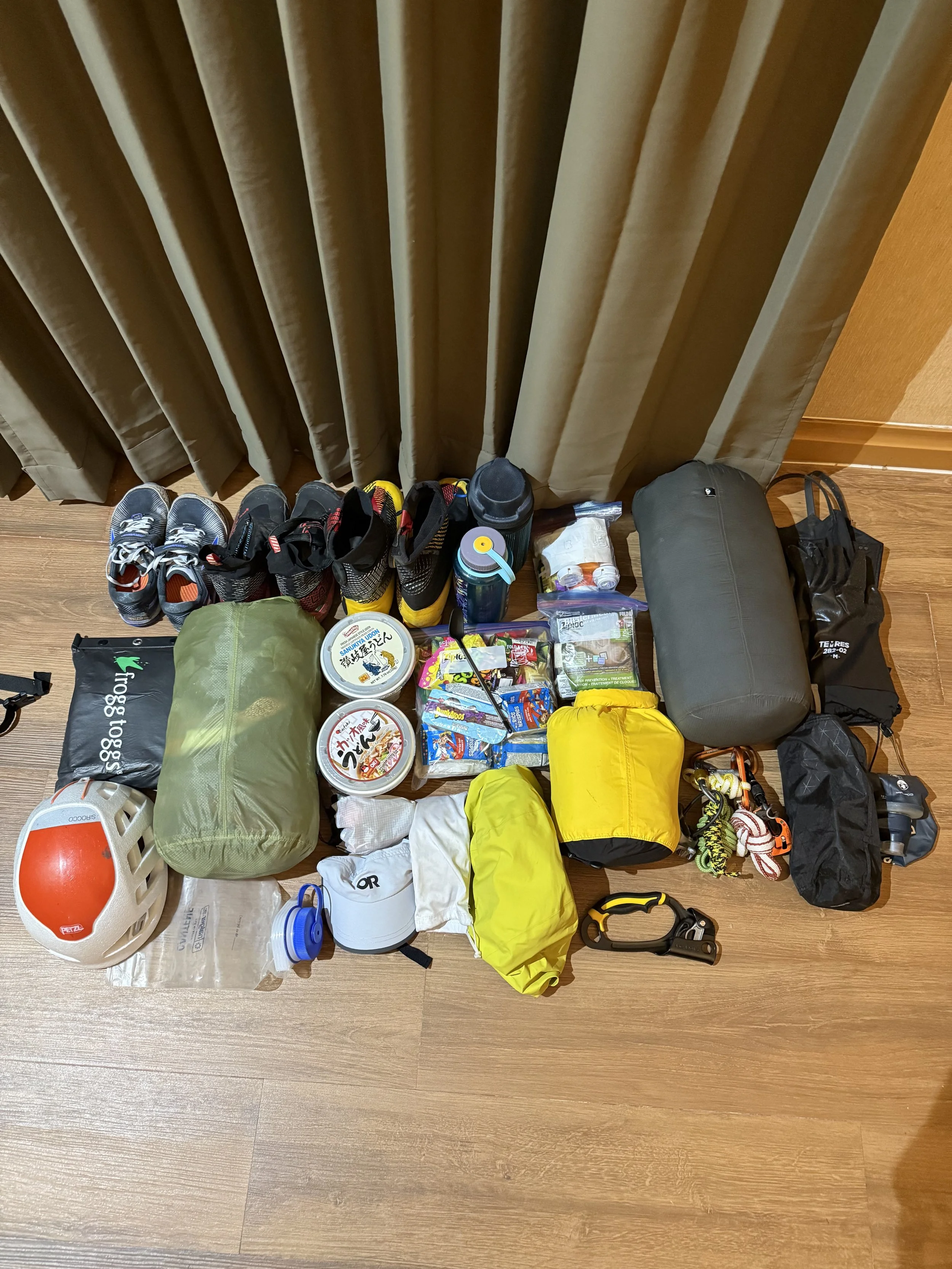 Assorted outdoor gear and supplies including shoes, water bottles, food, a helmet, and camping equipment arranged on the floor in front of curtains.