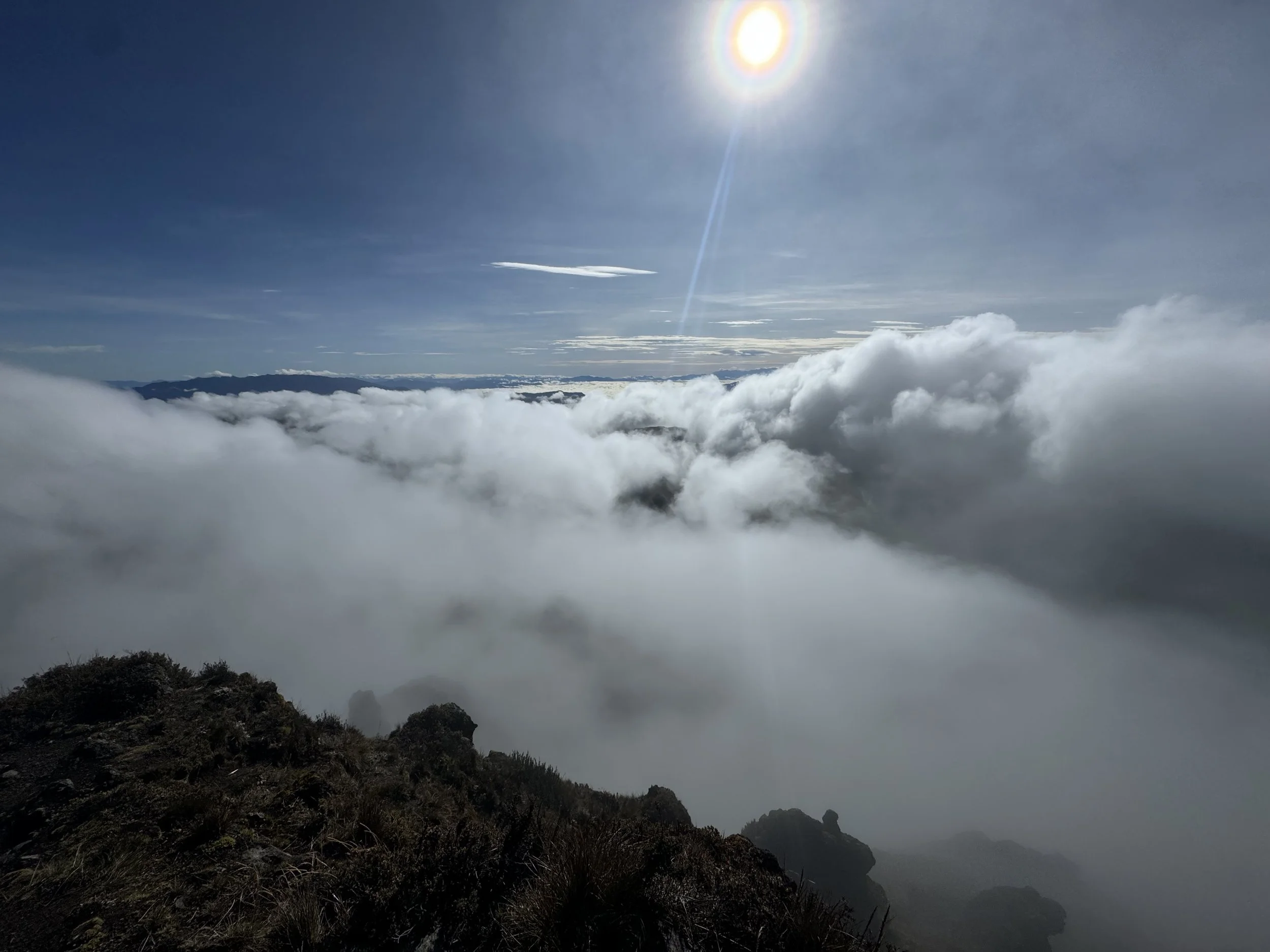 View from a mountain peak showing a sky with the sun, scattered clouds, and mountains below filled with fog.