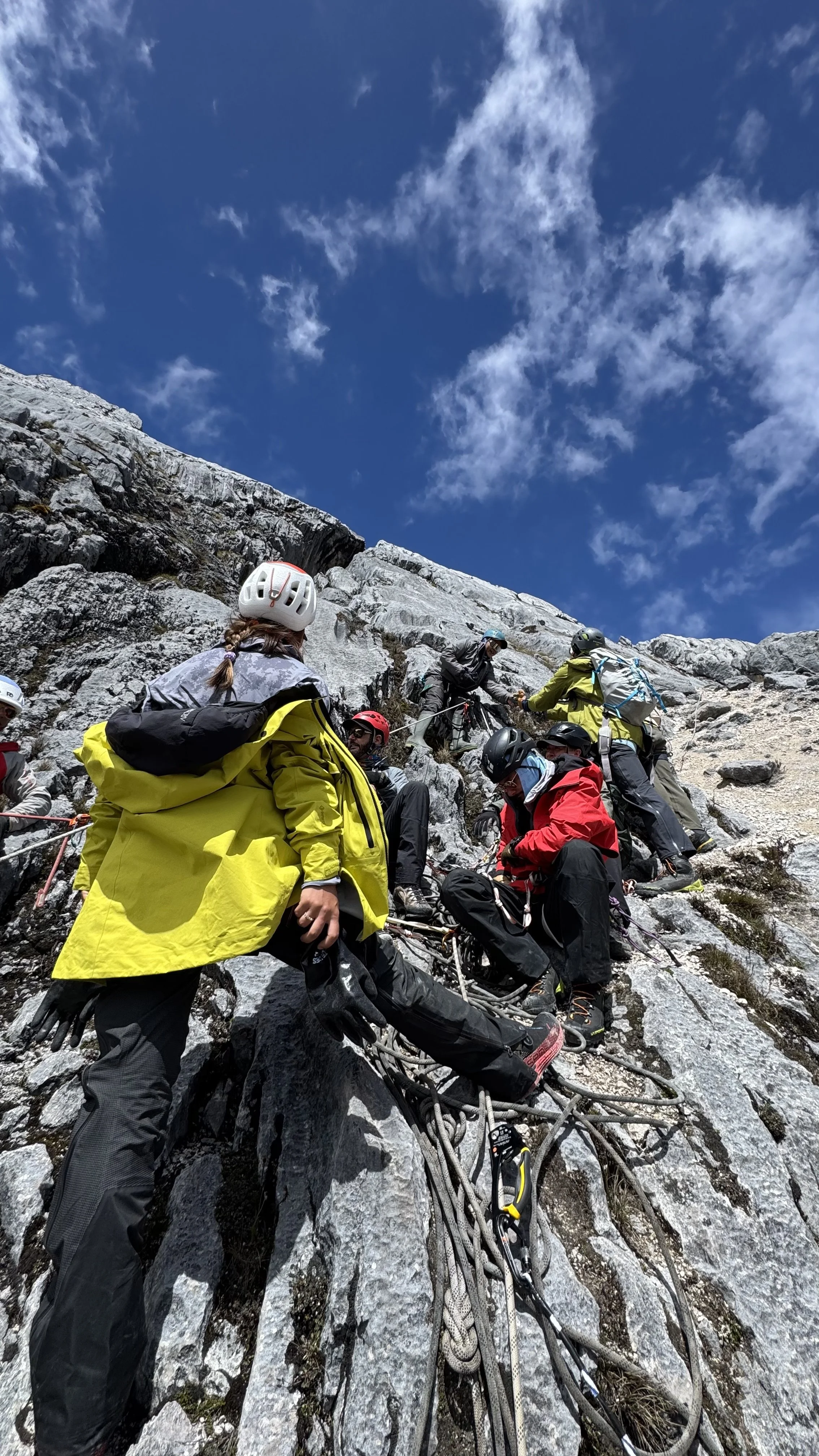 Group of climbers ascending a rocky mountain with mountain climbing gear and safety helmets under a partly cloudy sky.