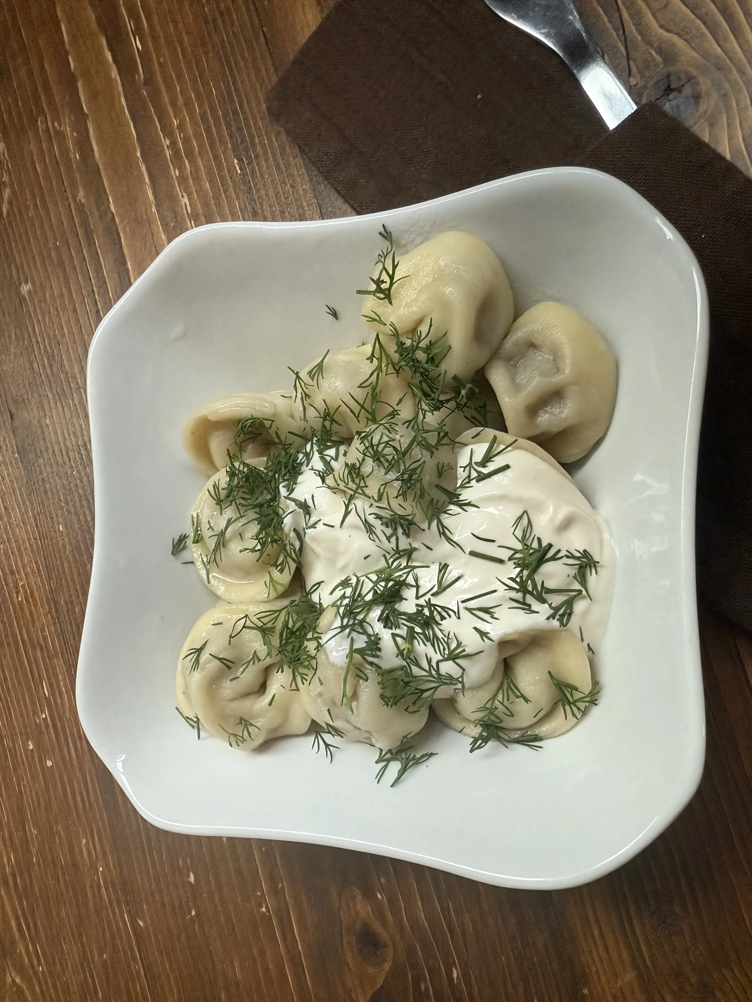 A white square bowl filled with dumplings topped with sour cream and garnished with fresh dill, placed on a wooden table.