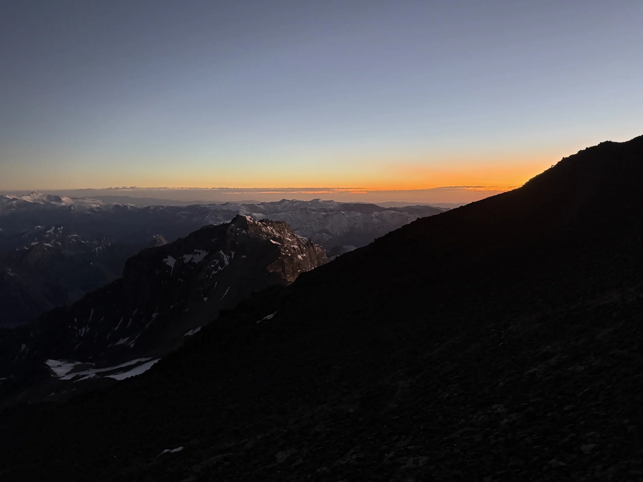 Sunset over snow-capped mountain peaks with a dark silhouette of a mountain in the foreground.