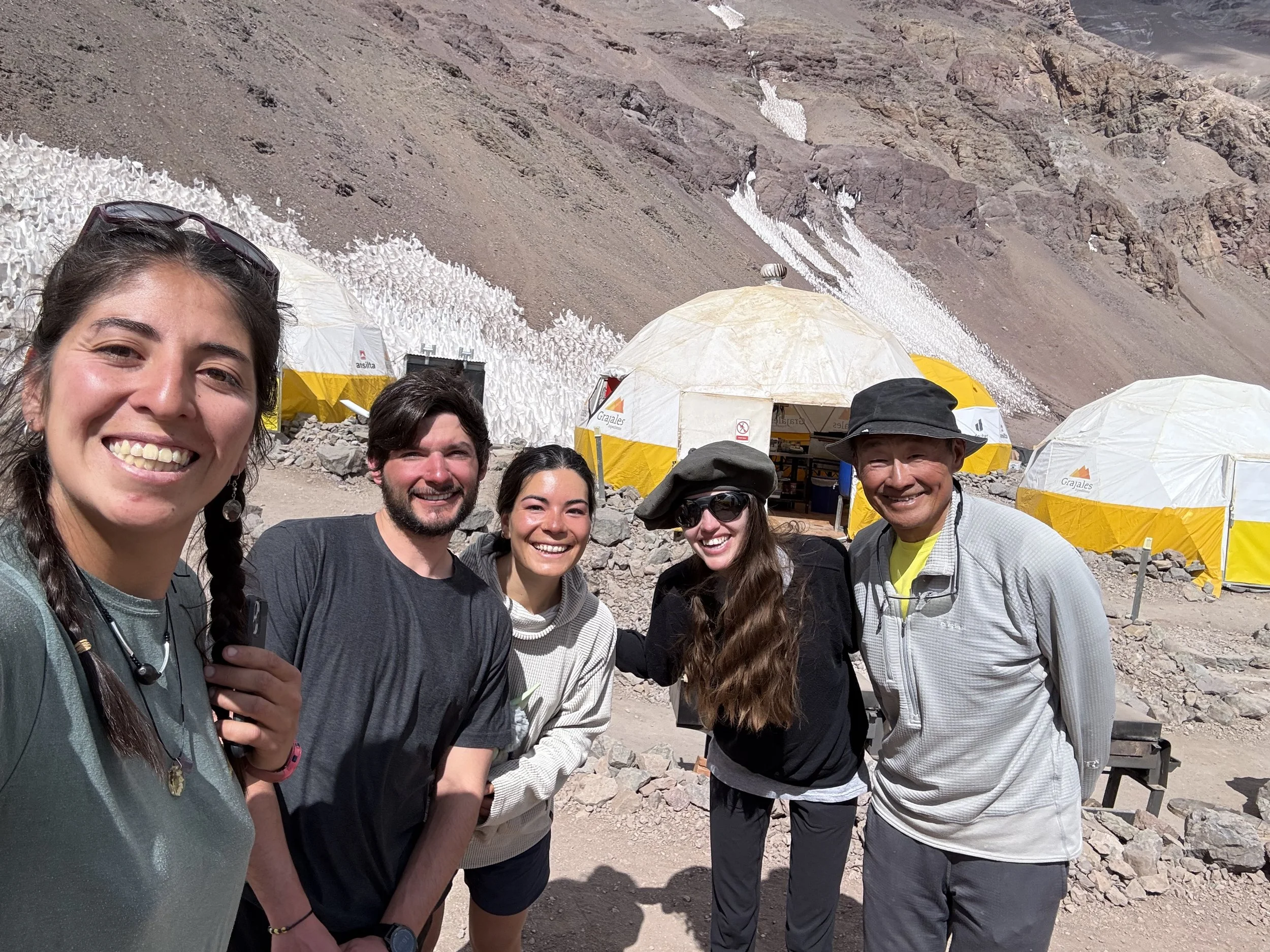 Group of five smiling people standing outdoors in front of mountain landscape with snow and tents.