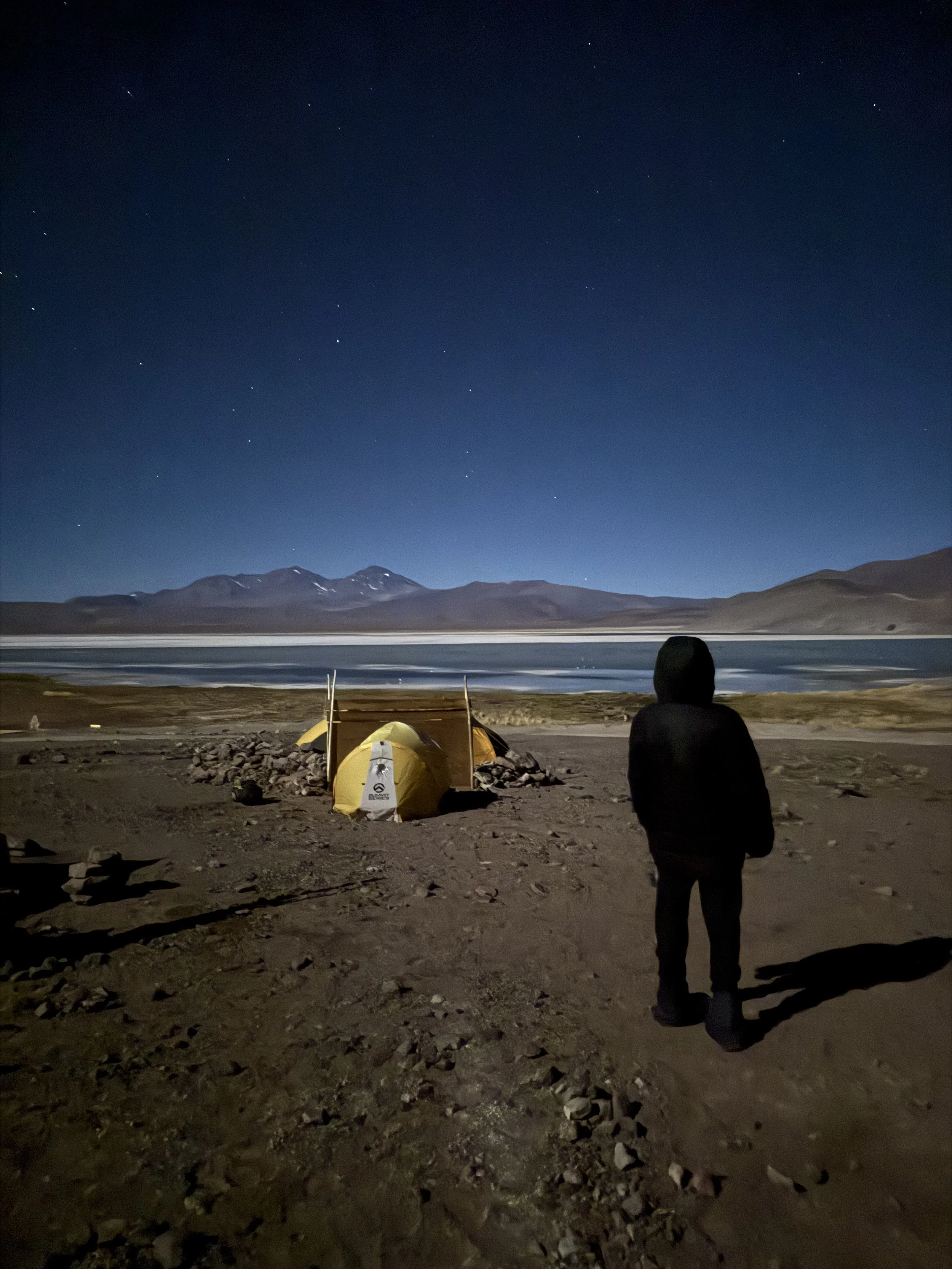 A person standing near a yellow tent at night, with a lake, mountains, and a starry sky in the background.