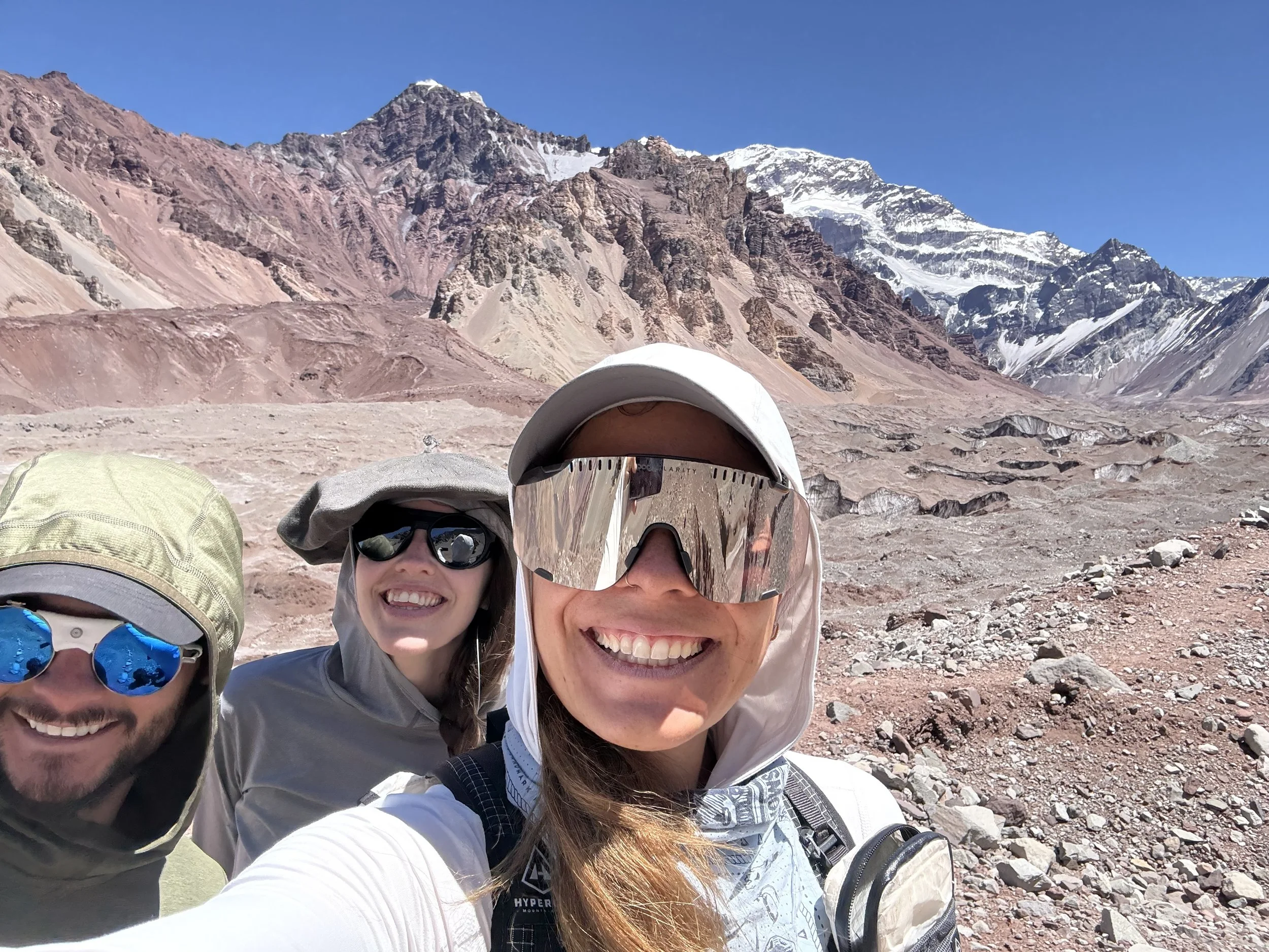 Three smiling hikers taking a selfie in a mountainous desert landscape with snow-capped peaks in the background.